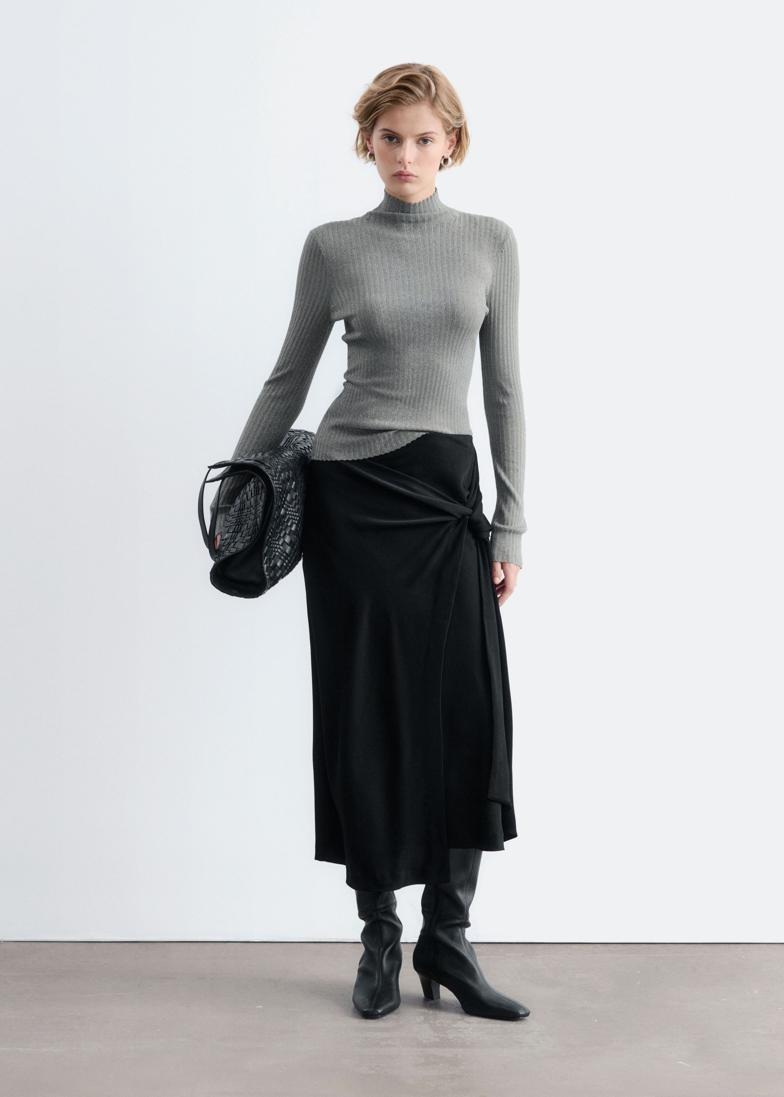 A gray, rib-knit mock neck top with long sleeves and a scalloped hem is worn by the model. The top is paired with a draped black midi skirt, black leather boots, and a woven black bag tucked under one arm.