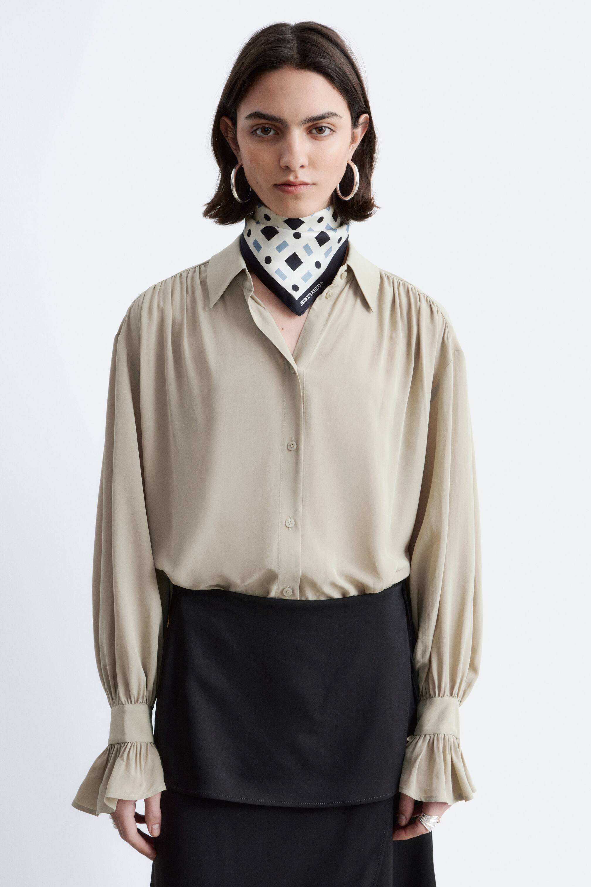 Agrandir l'image: Une blouse beige à manches longues présente une coupe décontractée, avec des épaules froncées, une fermeture boutonnée sur le devant avec un col en pointe, et des poignets à volants. Un foulard à motifs géométriques noir, blanc et bleu clair est noué autour de son col ouvert, et la blouse est rentrée dans une jupe noire.