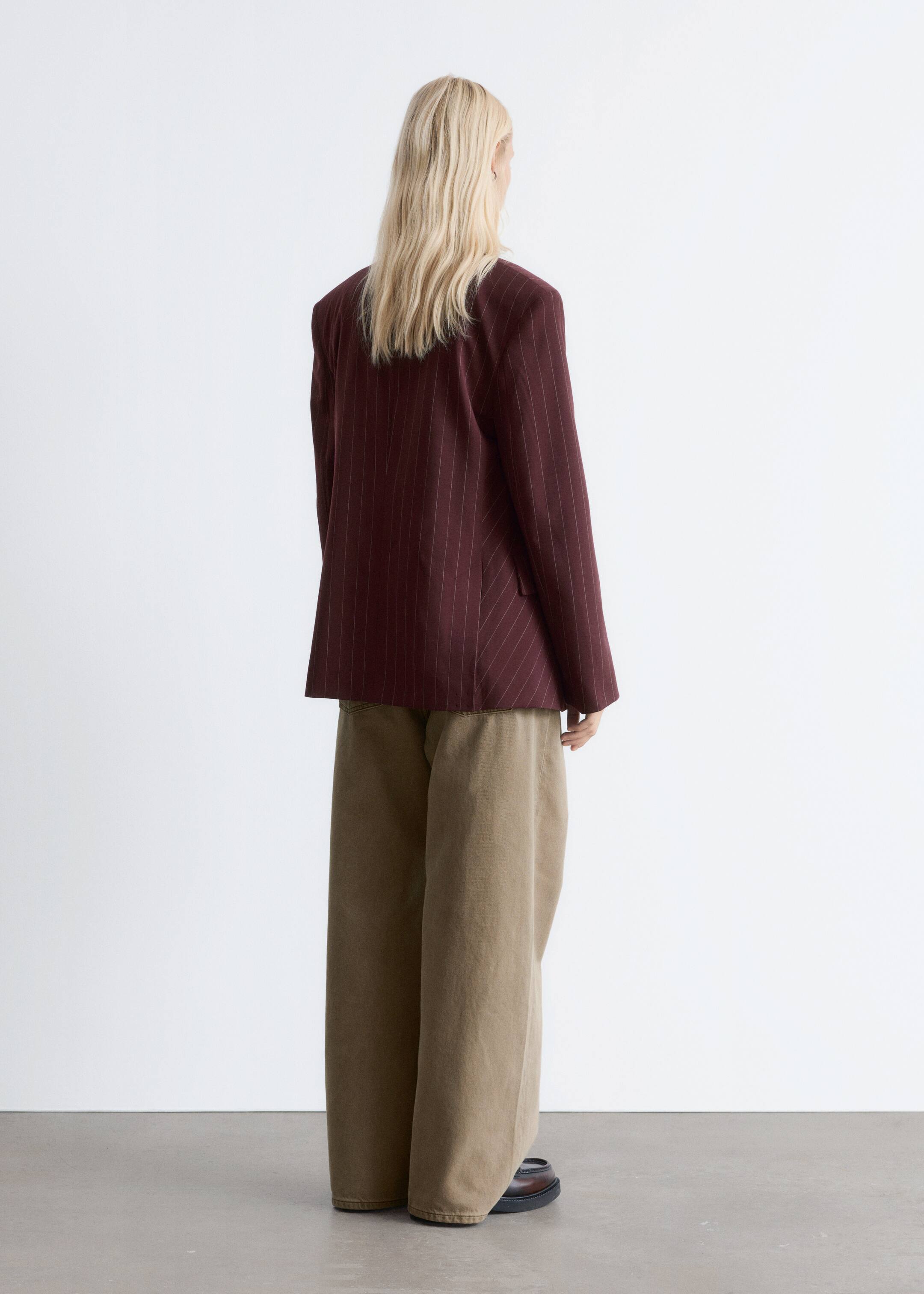 Agrandir l'image: Un blazer bordeaux à rayures est porté par le mannequin, qui se tient dos au spectateur. Le blazer présente des épaules structurées et est assorti à un pantalon large de couleur brun clair.