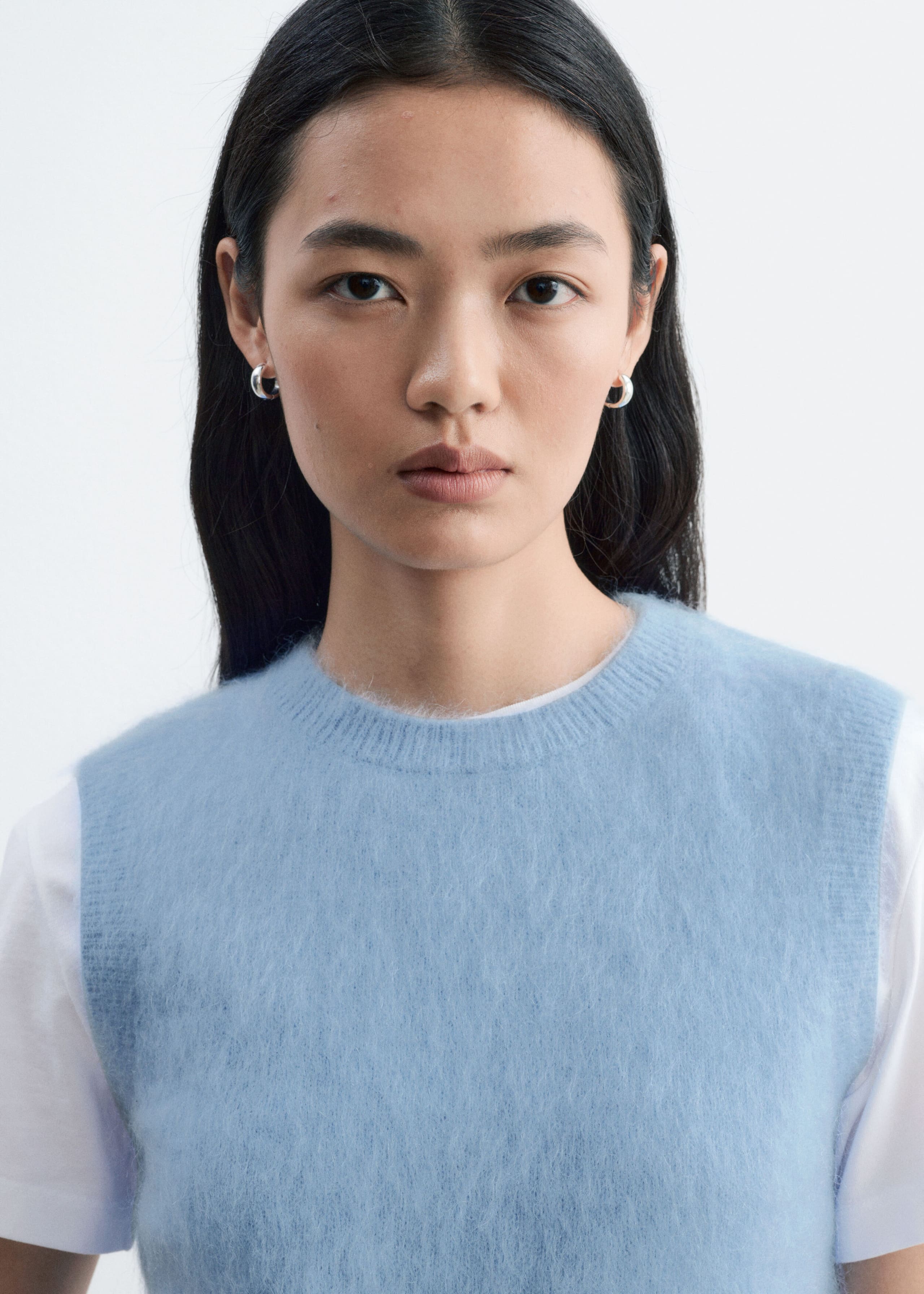 La modelo lleva un chaleco de mezcla de mohair de color azul claro, de punto, con una textura esponjosa y cuello redondo acanalado. El chaleco se superpone sobre una camiseta blanca de manga corta.
