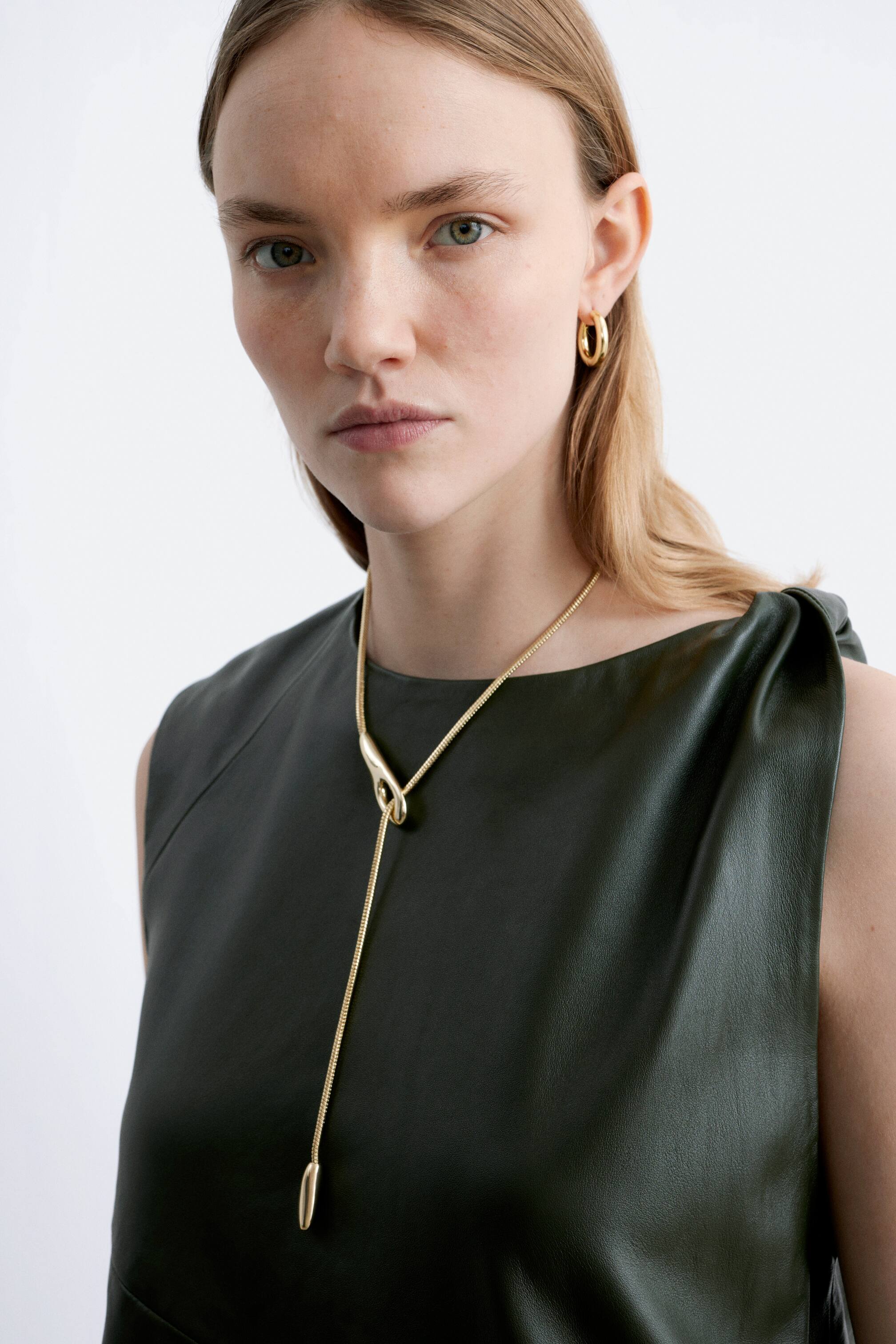 Grösseres Bild ansehen: Eine goldene Lariat-Halskette mit einer schlanken Kette, die durch ein poliertes, organisch geformtes zentrales Element verläuft. Die Kette erstreckt sich nach unten mit zwei verlängerten, kugelförmigen Enden, die auf einem dunkelgrünen ärmellosen Oberteil ruhen.