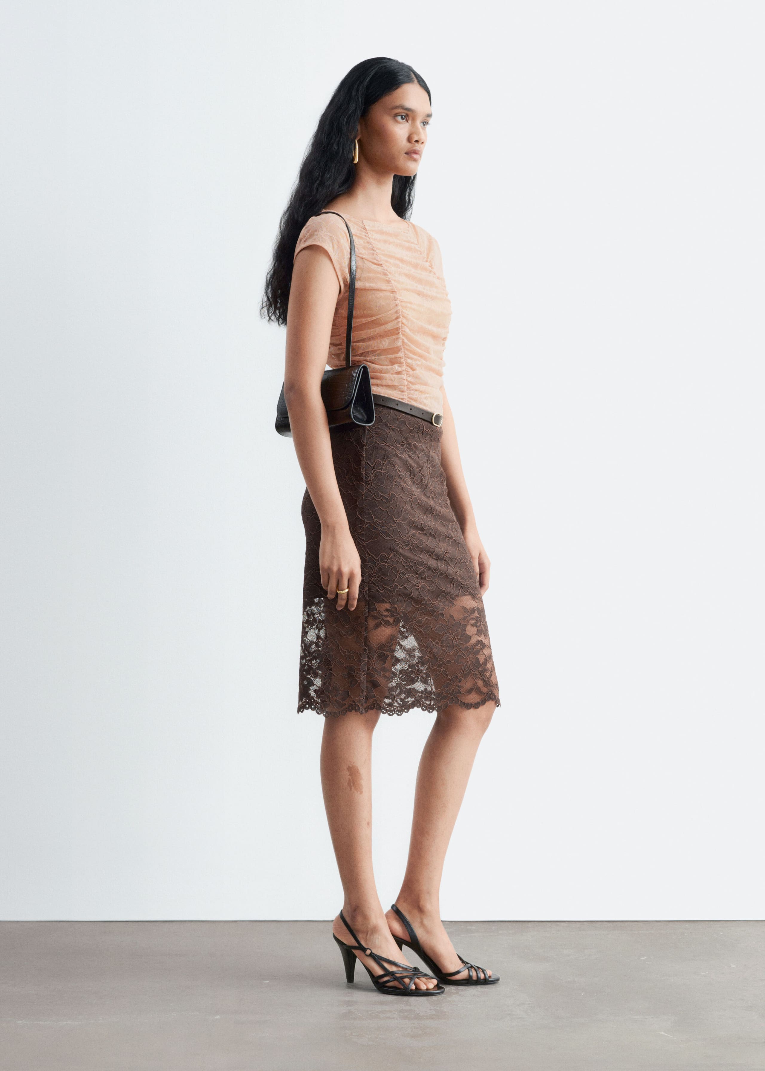 A peach-coloured ruched lace top featuring cap sleeves and a boat neckline is worn by the model. The top is tucked into a brown lace midi skirt and styled with a black shoulder bag.