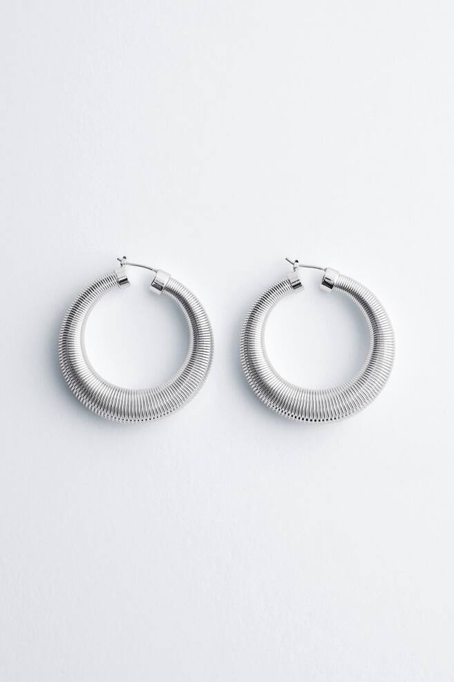 A pair of silver-toned hoop earrings lies flat and faces forward, showing a textured finish crafted from tightly coiled wire and featuring a hinge fastening at the top. - 1