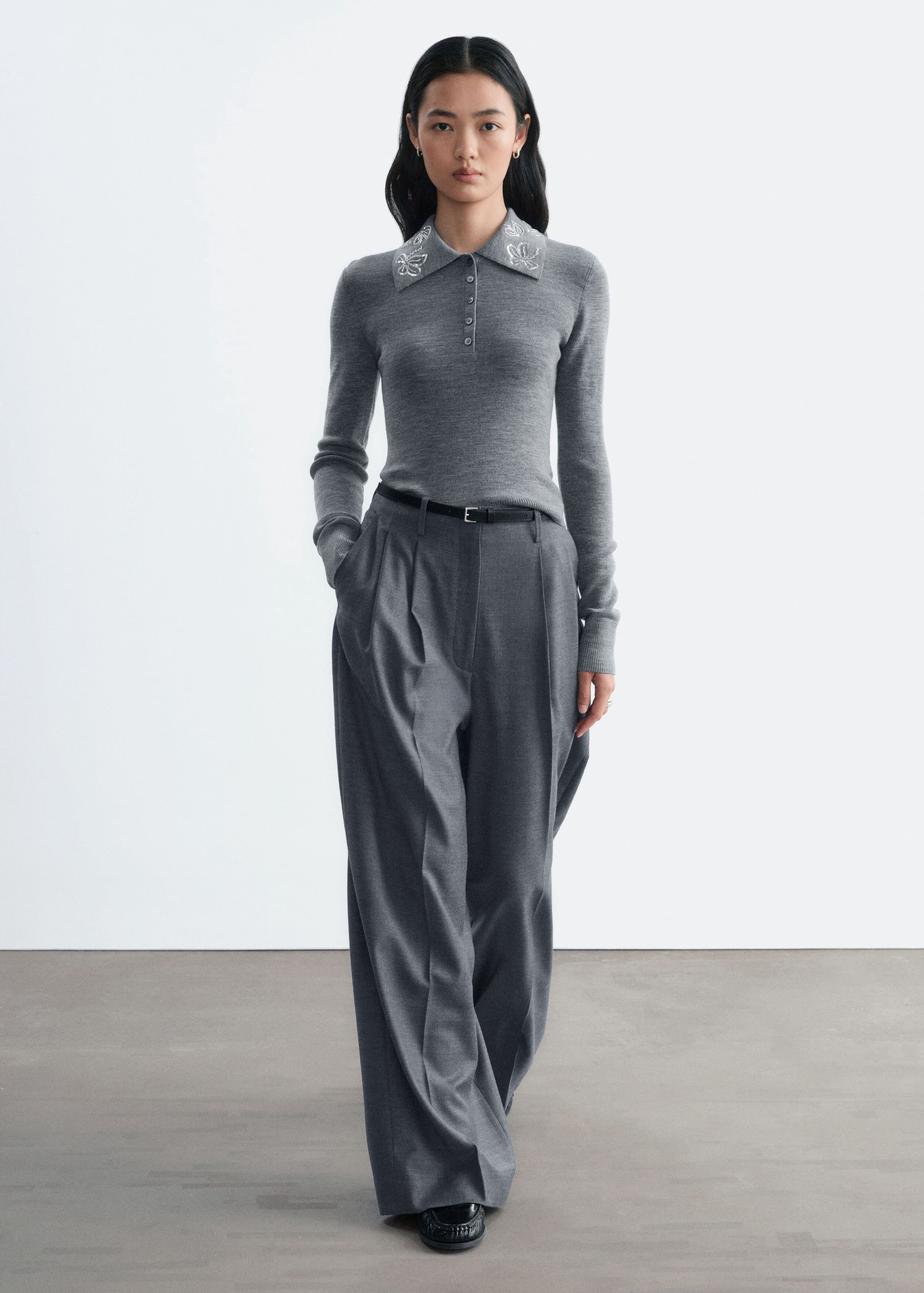 Ein schmal geschnittener, grauer Woll-Pullover mit Polo-Kragen, langen Ärmeln, geknöpfter Knopfleiste und weisser Blumenstickerei am Kragen wird vom Model getragen. Der Pullover ist in eine weite, plissierte graue Hose gesteckt und mit einem dünnen schwarzen Gürtel kombiniert.