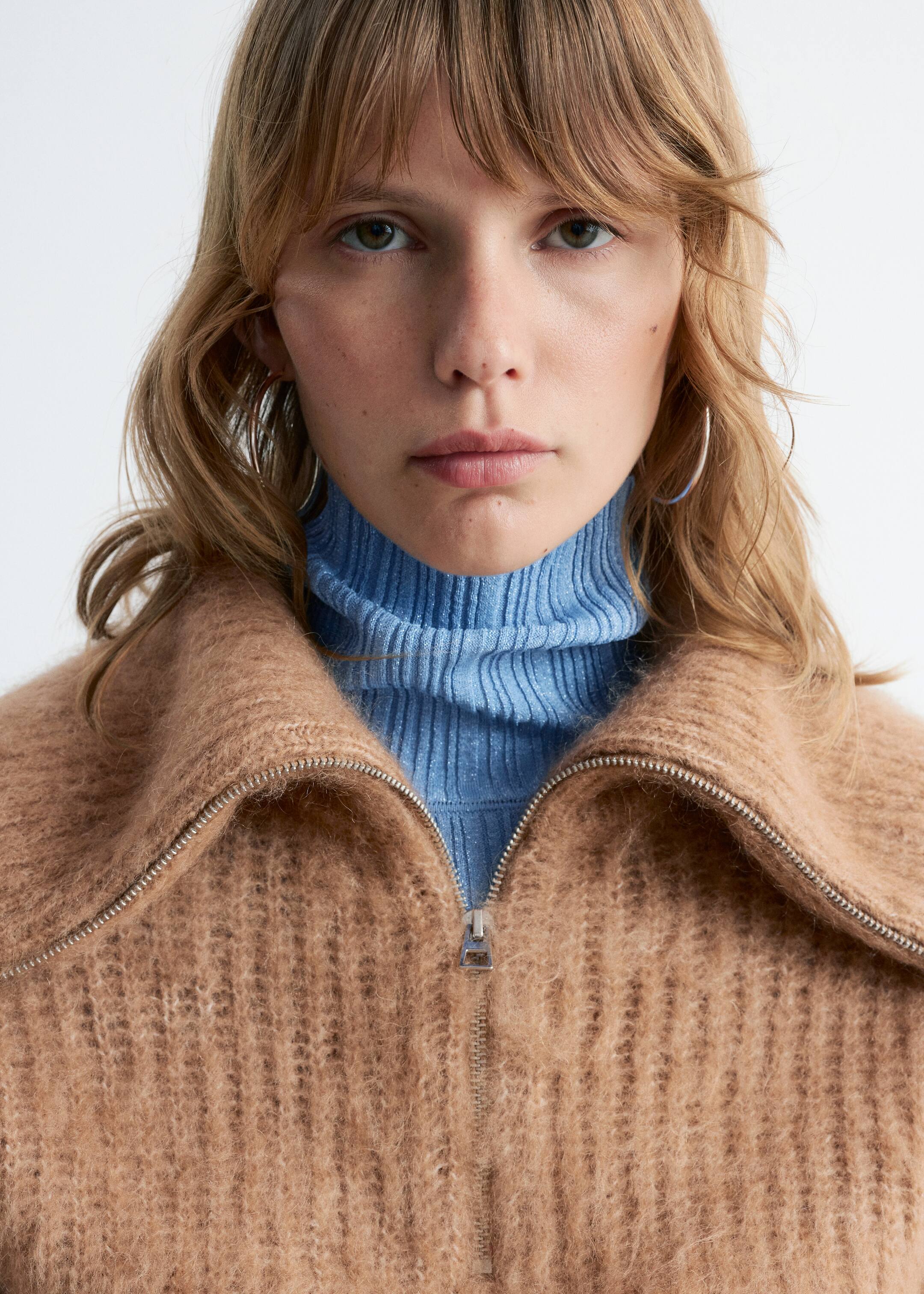 Grösseres Bild ansehen: Ein kamelfarbener, flauschiger Strickpullover mit halbem Reißverschluss wird getragen, der einen breiten, offenen Kragen und einen silberfarbenen Reißverschluss aufweist. Ein hellblauer gerippter Rollkragenpullover ist darunter sichtbar.