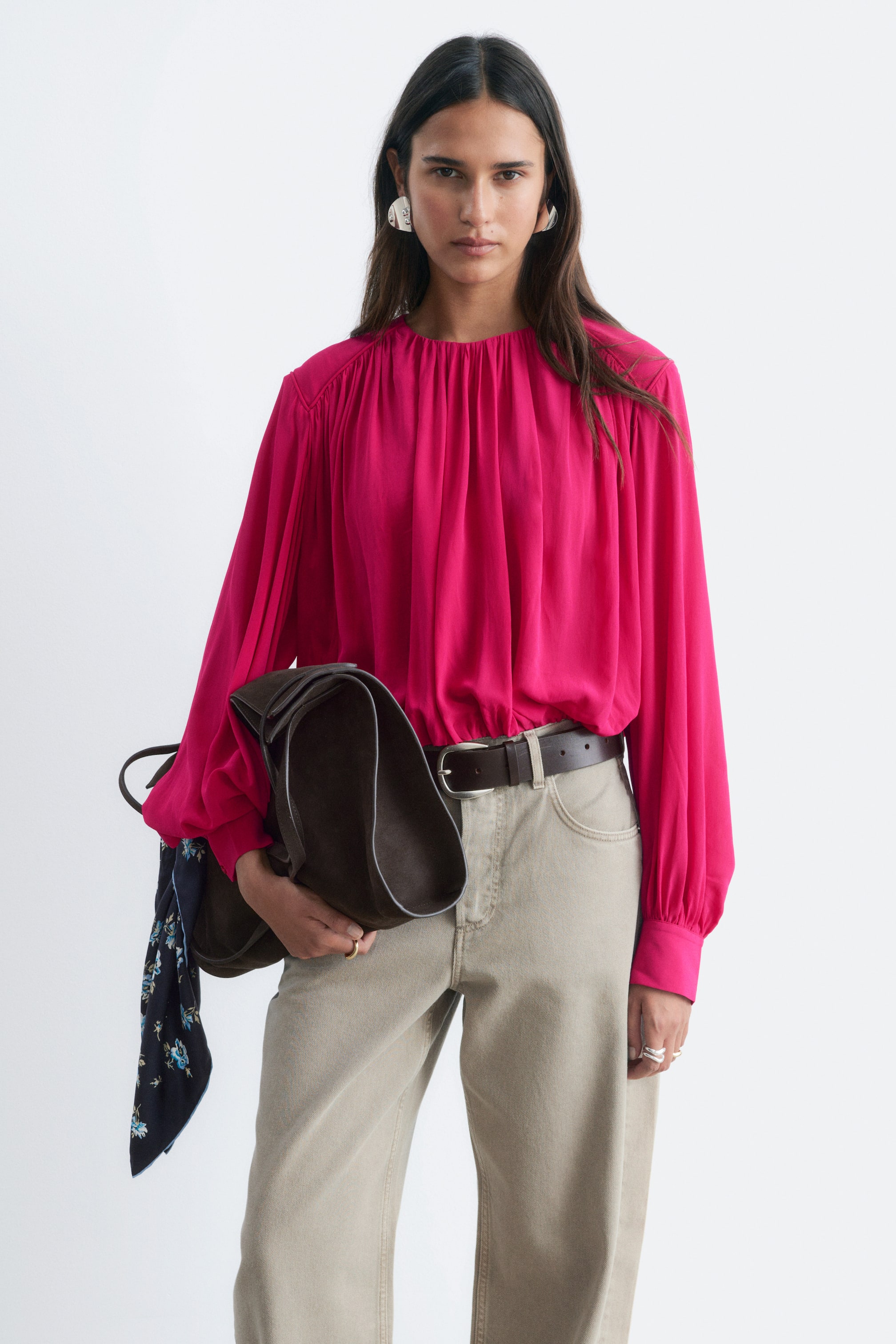 View larger image: The model is wearing a bright pink gathered blouse, featuring a gathered crew neckline and voluminous long sleeves with structured cuffs. The blouse is styled tucked into light beige trousers with a dark brown belt.