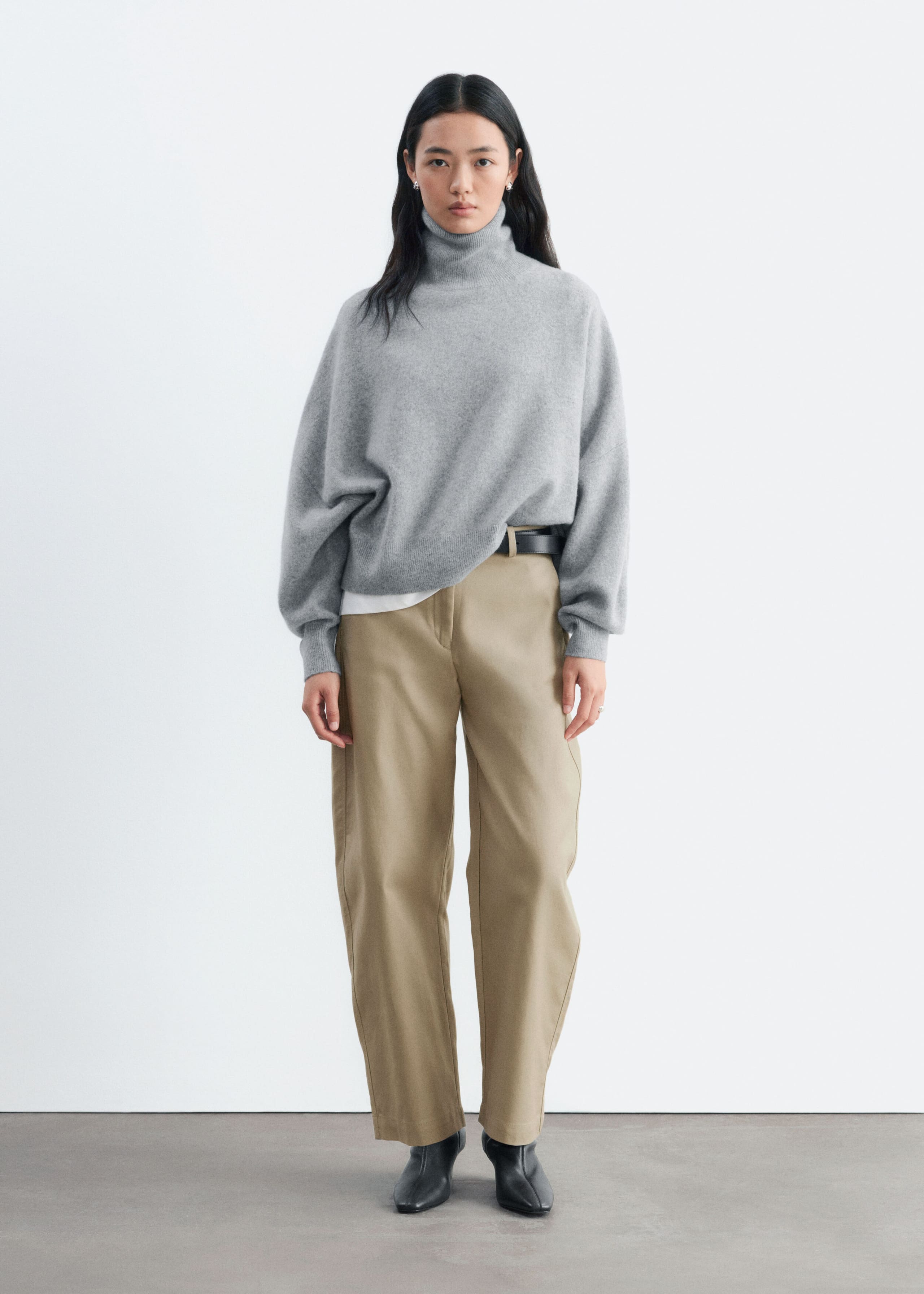 An oversized grey knit jumper with a folded roll neck and dropped shoulders is being worn by the model. The jumper is styled partially tucked into beige trousers at the waist, showing a white undershirt peeking out at the hem.
