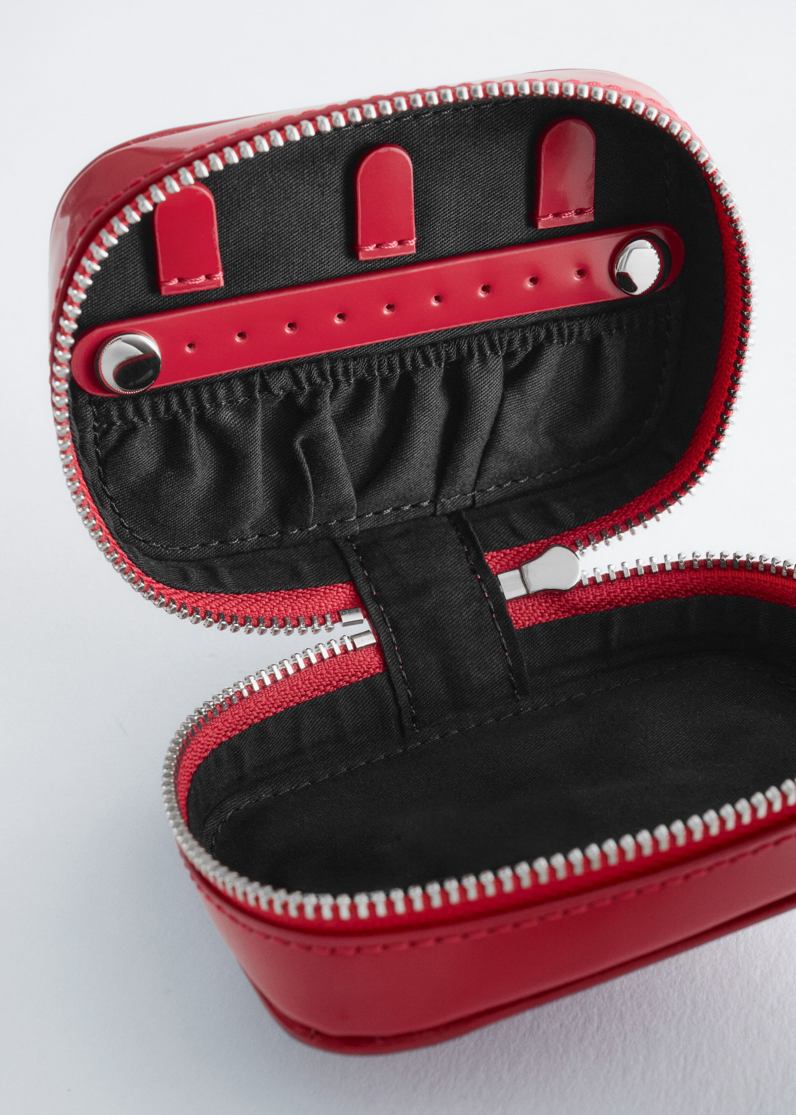 An open red jewellery case is shown from the front, displaying its black-lined interior, featuring three red snap-button loops, a perforated earring strap with silver snaps, and an elasticated slip pocket on the upper lid. A silver-tone metal zip runs along the edges of the red outer shell, opening to reveal an empty main compartment.