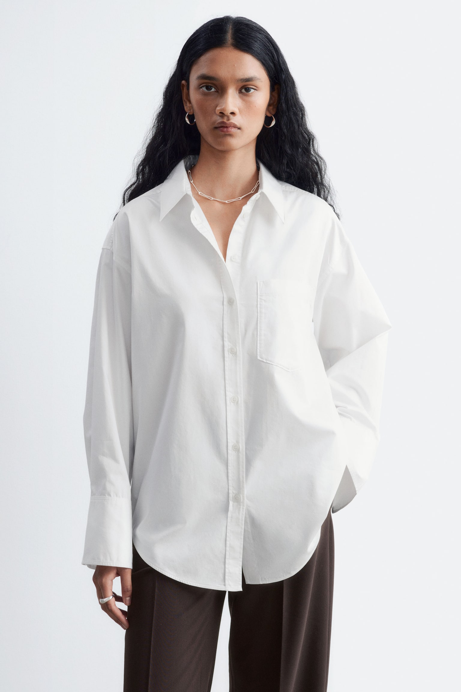 Le mannequin porte une chemise blanche en coton Ã  coupe ample, dotÃ©e dâun col pointu, dâune poche plaquÃ©e sur la poitrine et de manches longues avec larges poignets. La chemise est portÃ©e sortie sur un pantalon marron foncÃ©. - 1