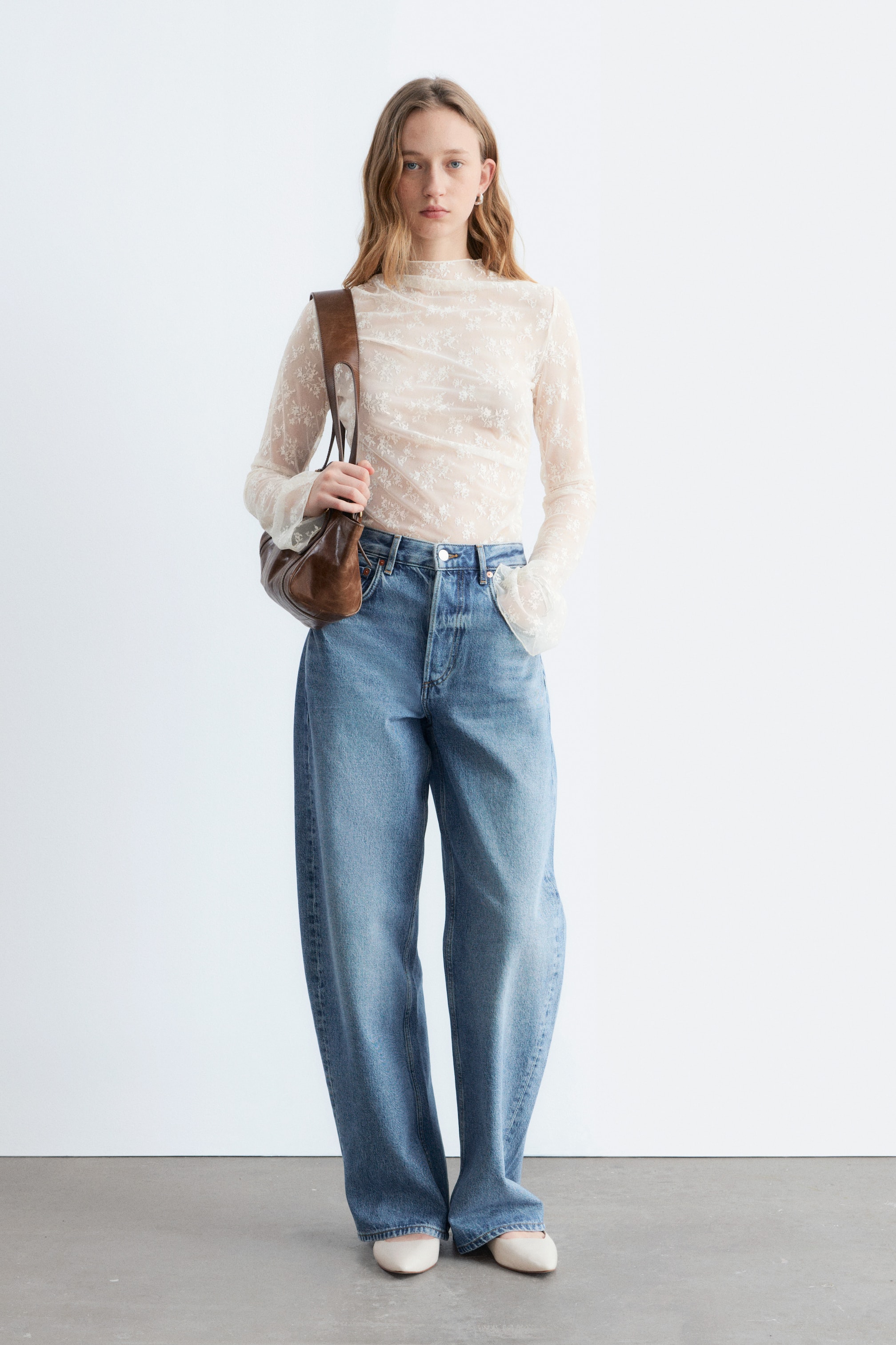 Agrandir l'image: Un haut léger crème transparent, orné d'un délicat motif floral, avec un col montant et des manches longues, est porté rentré dans un jean bleu taille haute à jambes larges, complété par des ballerines à bout pointu crème.