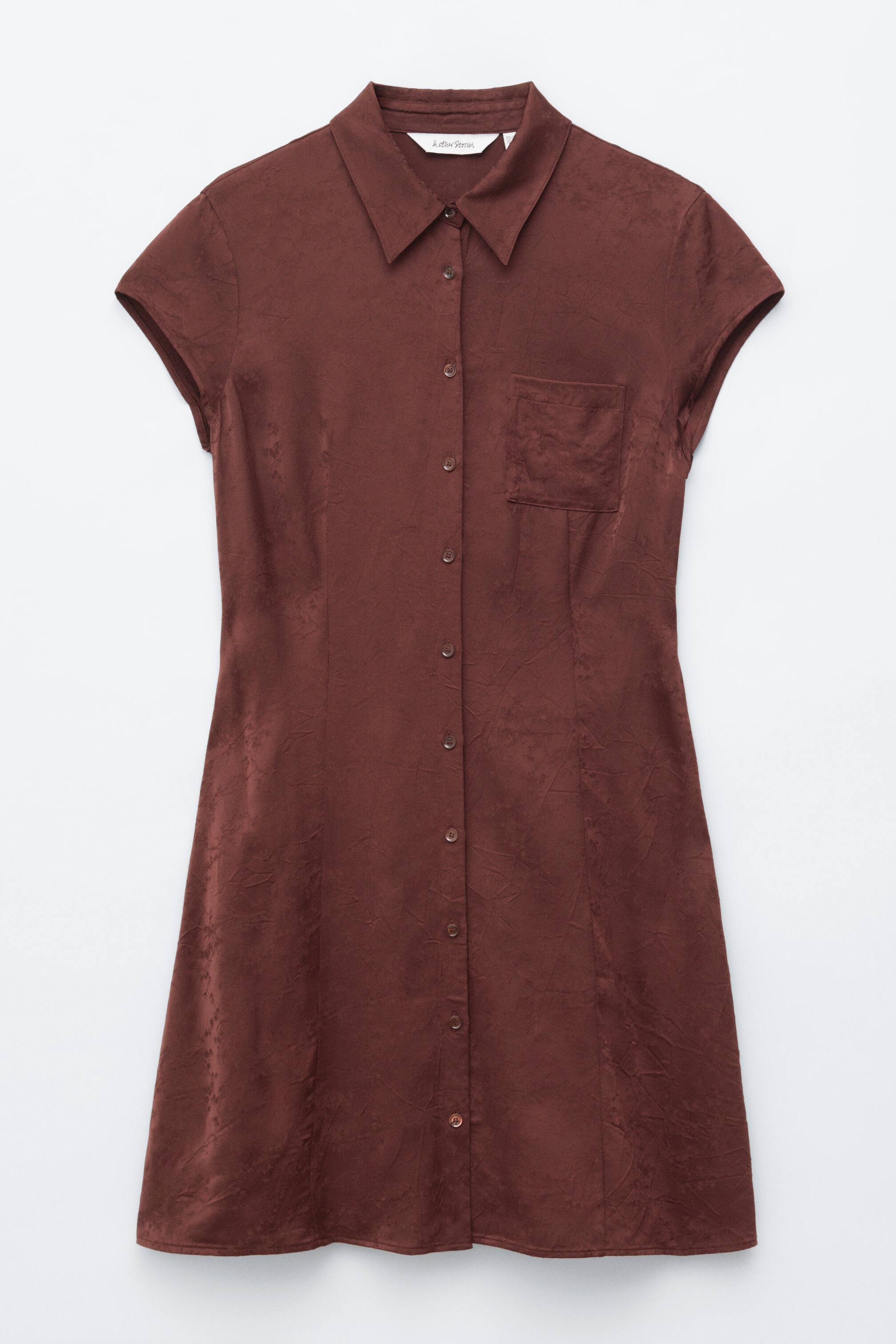 Visa större bild: En brun miniskjortklänning visar sin framsida, med en skjortkrage, en full knappslå och korta ärmar. En enda bröstficka syns på vänster sida, och ett subtilt texturerat mönster framträder i hela tyget.