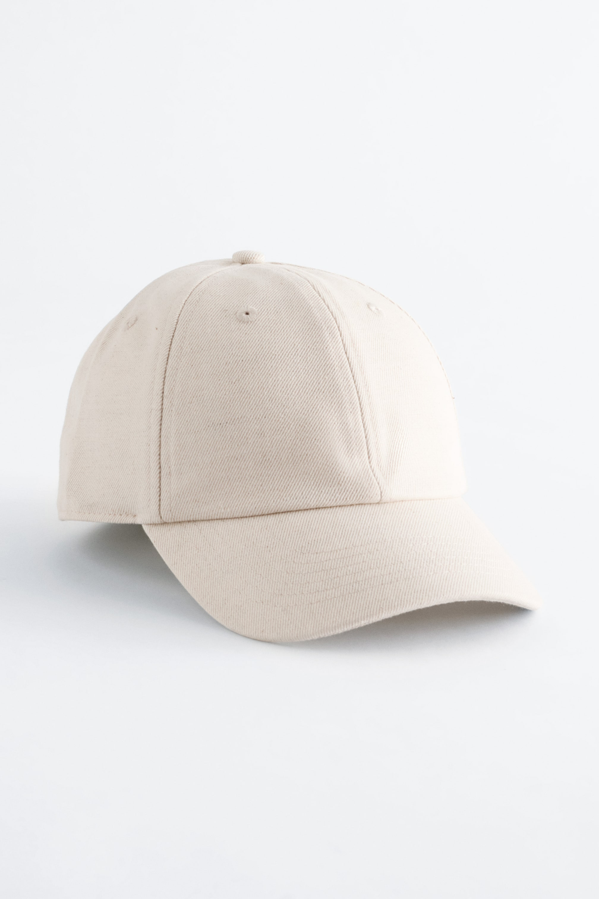 Ingrandisci l'immagine: Un berretto da baseball in cotone-lino color beige chiaro presenta il suo fronte e lato, inclinato leggermente verso destra, ed è caratterizzato da una visiera curva, un bottone superiore e due occhielli di ventilazione sui pannelli frontali.