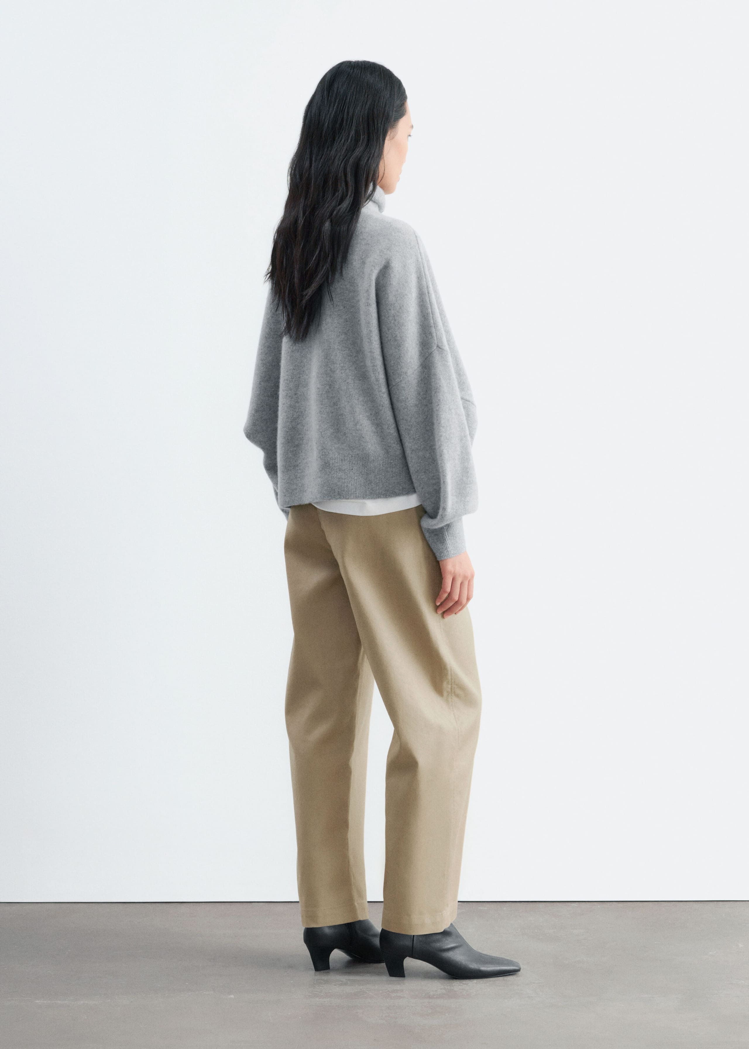 An oversized grey knit jumper featuring dropped shoulders and a high collar is worn by the model, shown from a three-quarter back view. The jumper is styled with a white top layered underneath, beige wide-leg trousers, and black leather boots.
