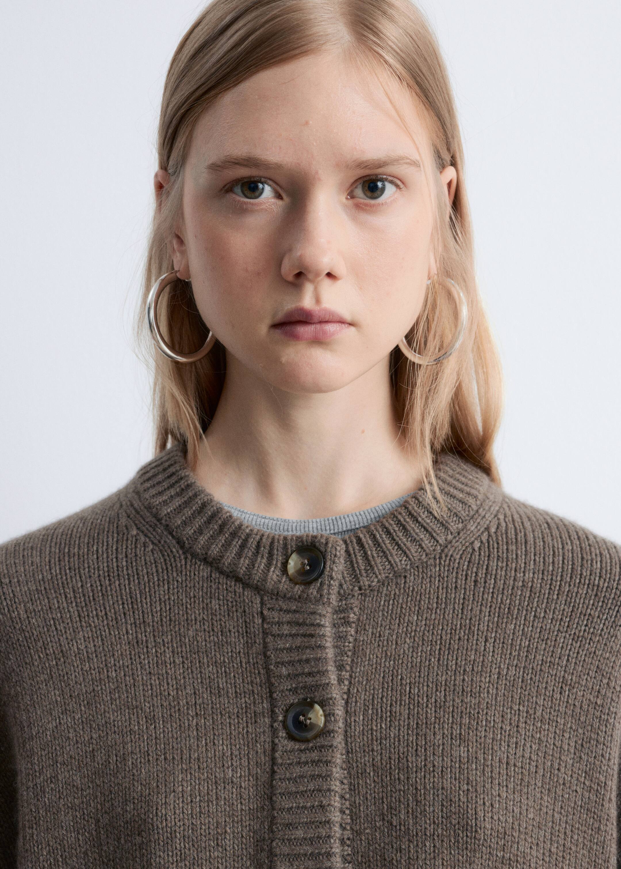 Agrandir l'image: Un cardigan tricoté marron est porté face avant, avec un col rond côtelé et une patte de boutonnage côtelée à l'avant avec deux boutons visibles en écaille de tortue foncée. Un haut à col rond gris est visible sous le cardigan.
