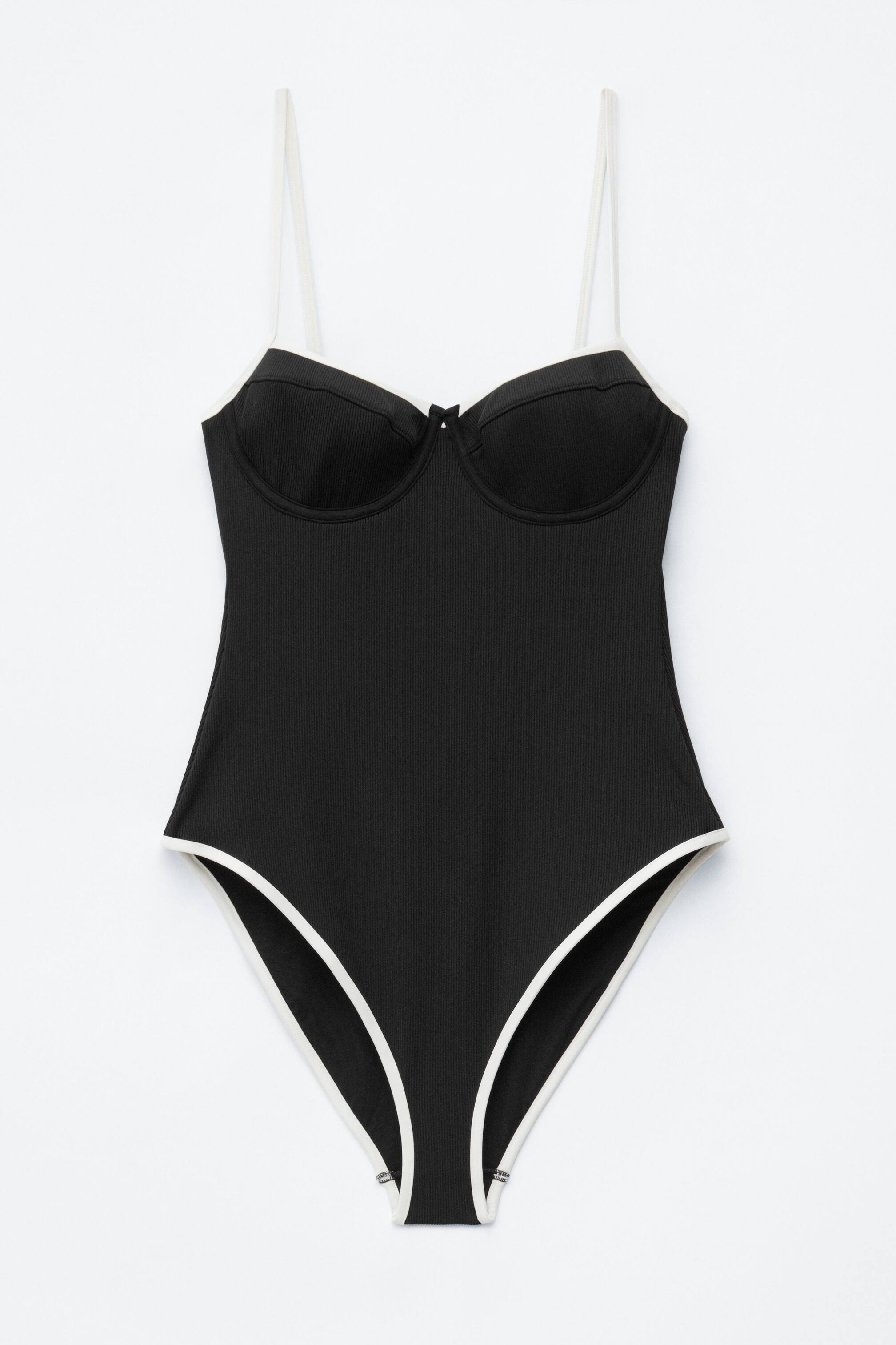 Ver imagen más grande: Un bañador negro de canalé presenta su parte frontal, con copas moldeadas con costuras pespunteadas y un pequeño detalle de lazo en el centro. Un ribete blanco en contraste perfila el escote, los tirantes ajustables y las aberturas de pierna de corte alto.