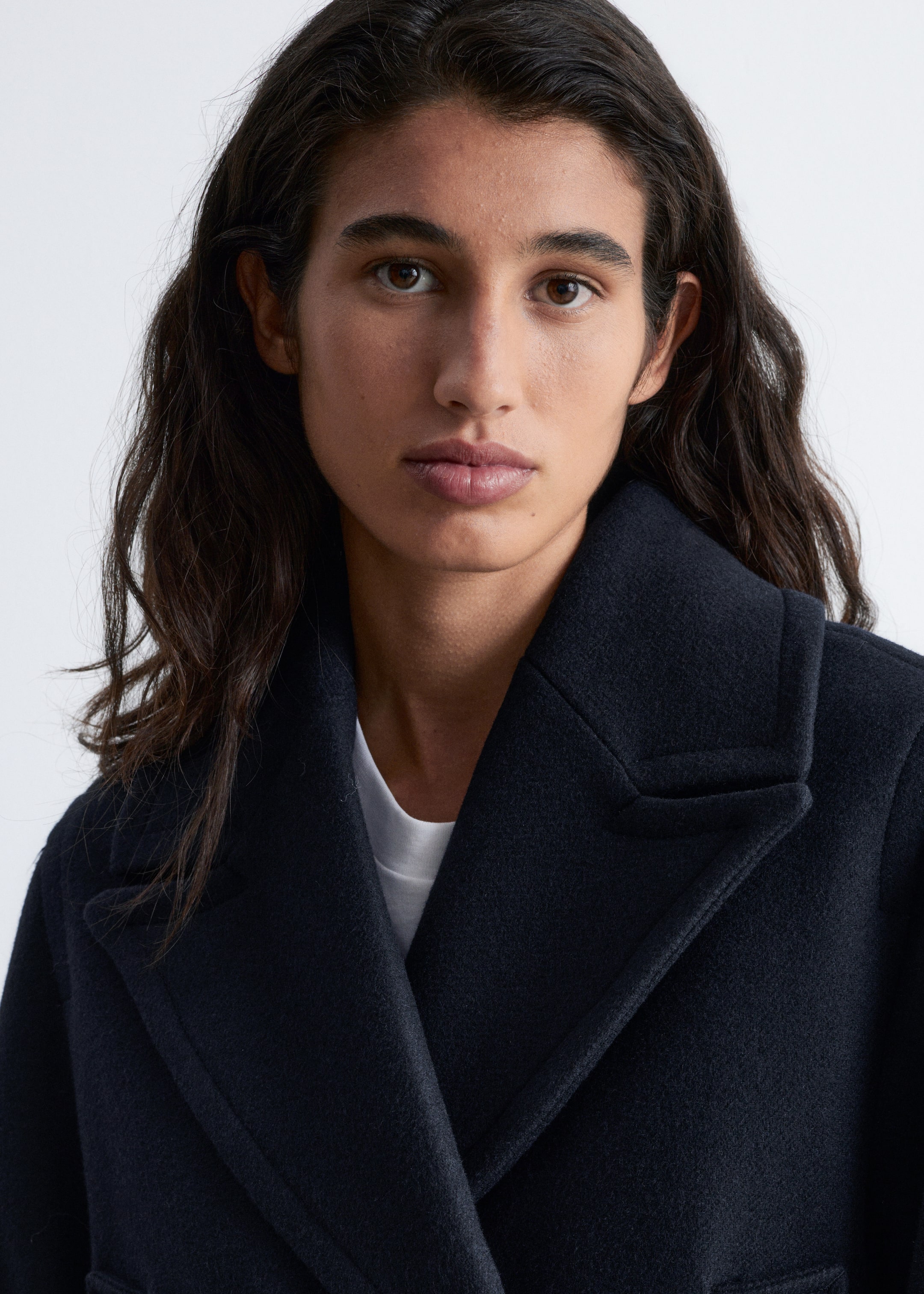 View larger image: A navy blue coat is being worn, featuring wide peak lapels and a structured, overlapping front, presented from a slightly angled front view. A white t-shirt is being worn underneath the coat, visible through the open front.