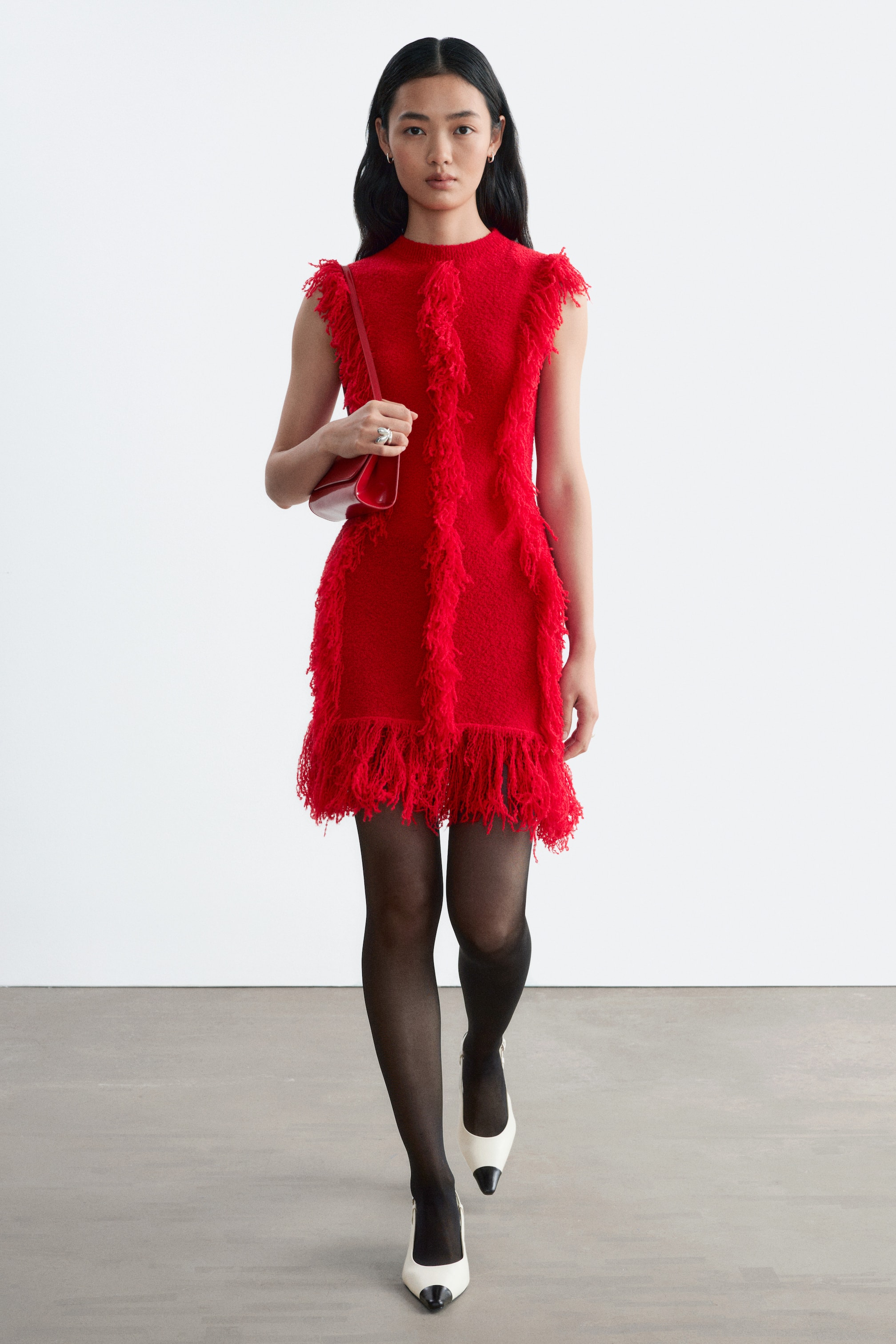 Ver imagen más grande: La modelo lleva un vestido de punto sin mangas de color rojo vivo, con filas verticales de flecos texturizados y bajo con flecos. El vestido de corte entallado se combina con medias negras transparentes, zapatos de tacón blancos de punta con punteras negras y un pequeño bolso de hombro rojo llevado bajo el brazo.
