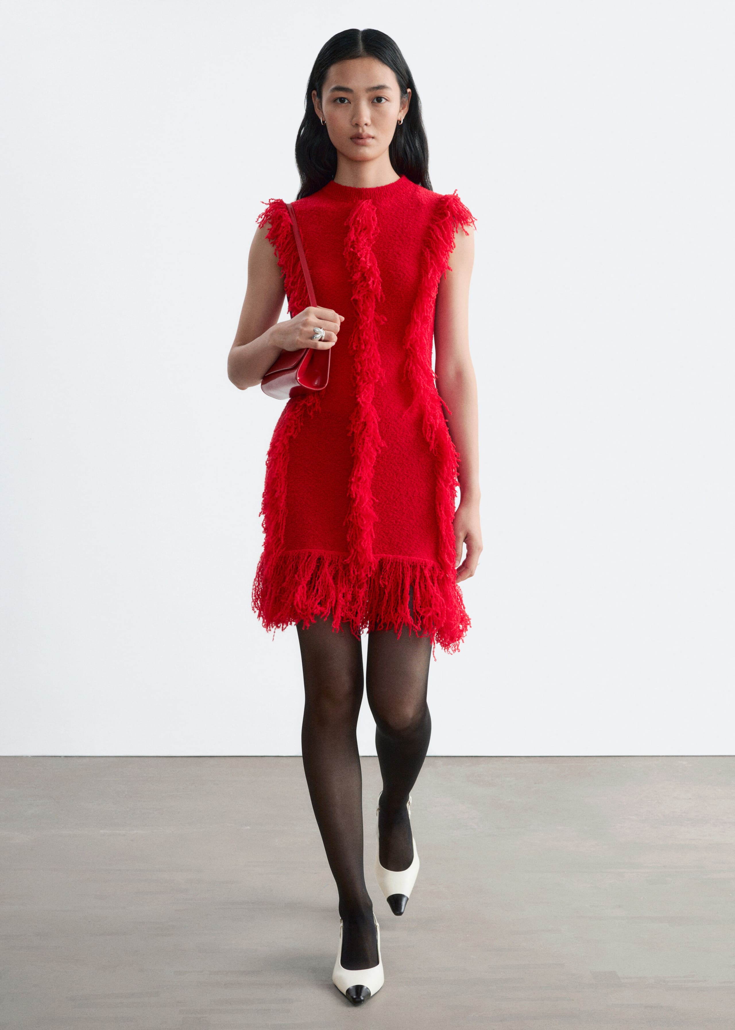 A bright red, sleeveless knit dress featuring vertical rows of textured fringe and a fringed hem is being worn by the model. The slim-fit dress is styled with sheer black tights, white pointed-toe heels with black caps, and a small red shoulder bag carried under the arm.