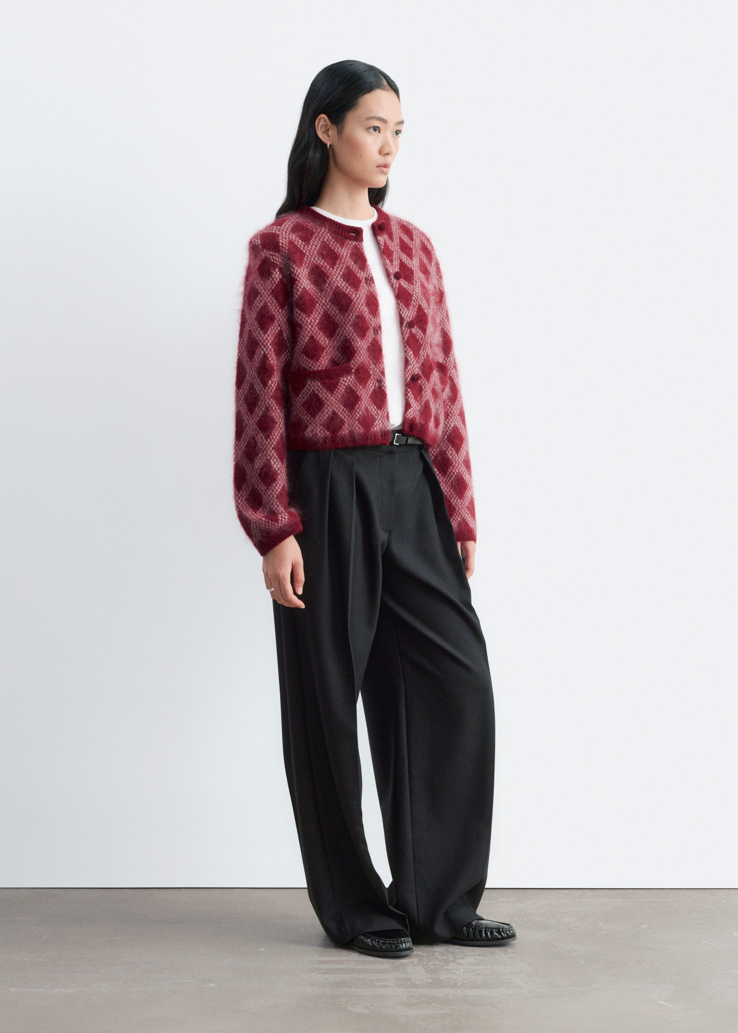 A red and cream diamond-patterned fuzzy knit cardigan is being worn by the model, layered over a white top and wide-leg black trousers. The cardigan features structured shoulders, long sleeves with ribbed cuffs, and a ribbed hem.