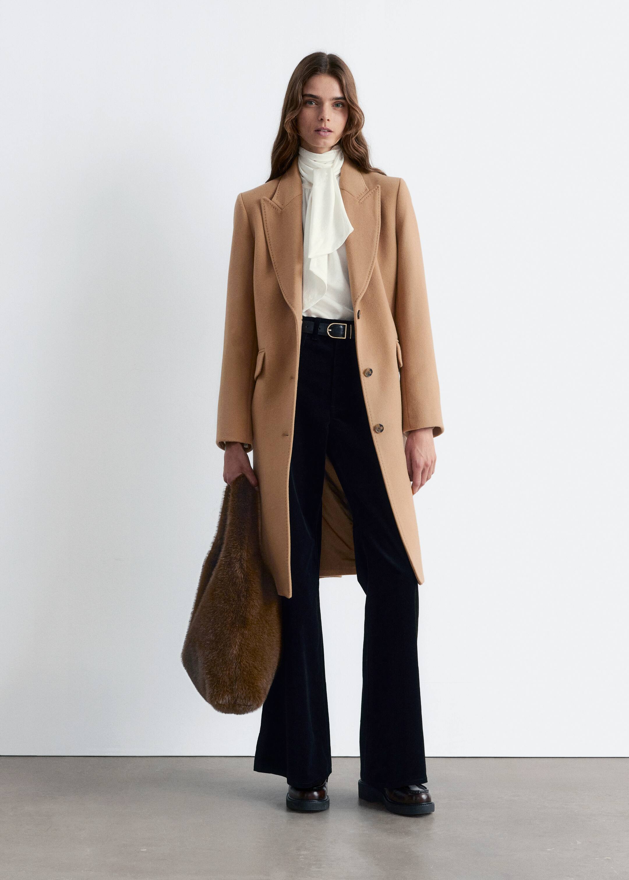 View larger image: A long, camel-colored coat is being presented head-on, worn open over a white high-neck blouse and black wide-leg trousers. It is featuring structured shoulders, notch lapels, a single-breasted front with two visible buttons, and two flap pockets, extending to mid-calf.