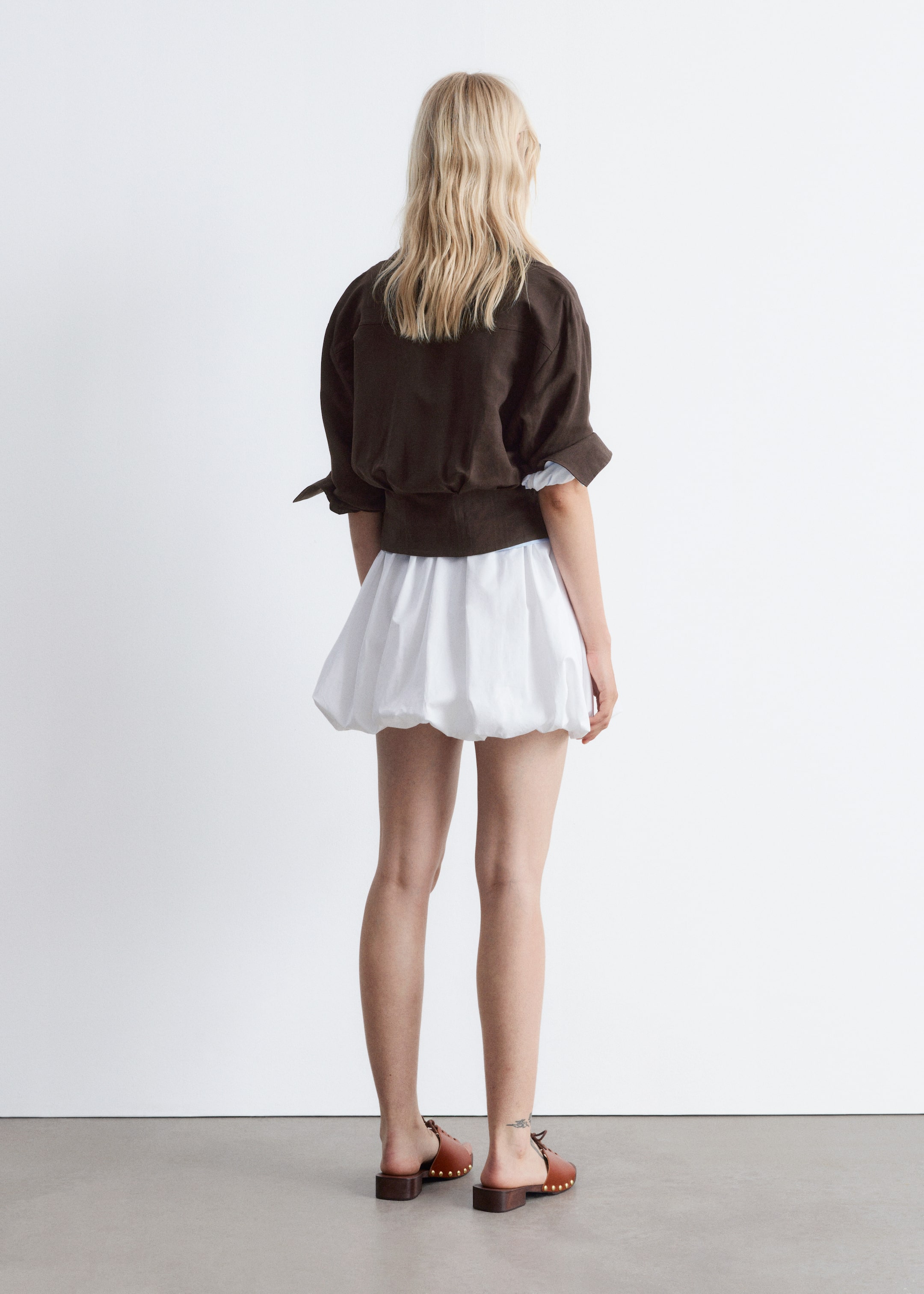 Größeres Bild anzeigen: Ein dunkelbraunes Langarmhemd wird von dem Modell getragen, das mit dem Rücken zum Betrachter steht. Das Hemd hat eine geraffte Taille, die einen Blouson-Effekt erzeugt, und die Ärmel sind hochgekrempelt, wodurch ein weißes Unterhemd sichtbar wird.