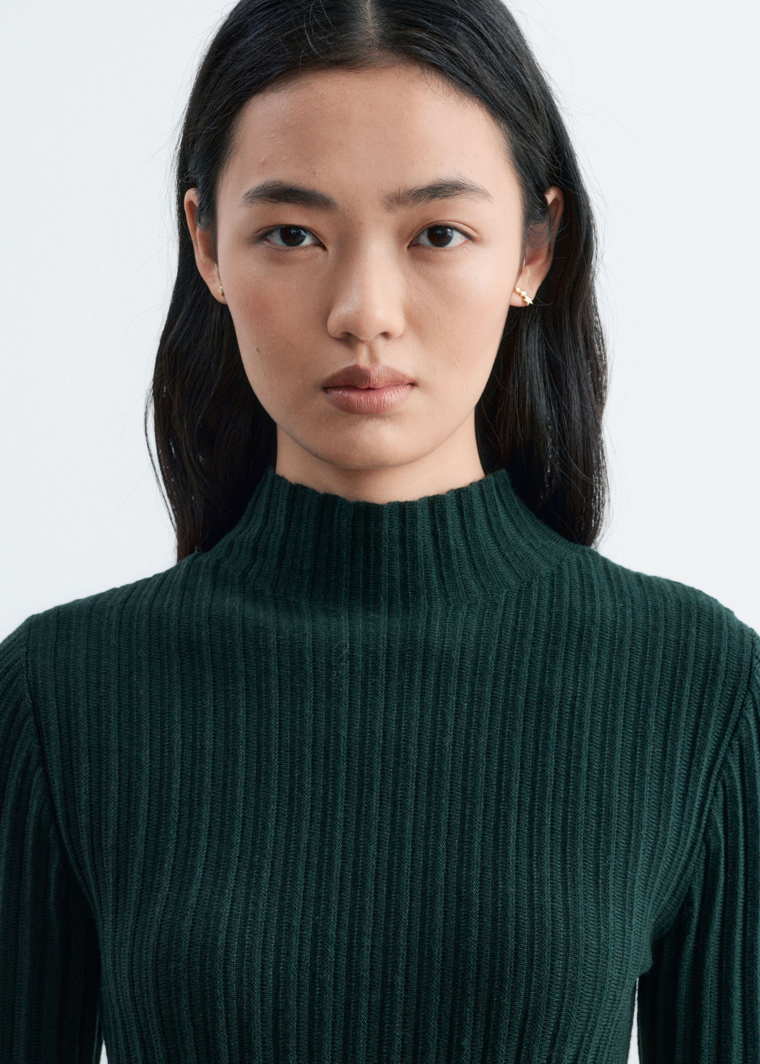 A dark green, ribbed knitted top featuring a roll neck is being worn by the model in a close-up view.