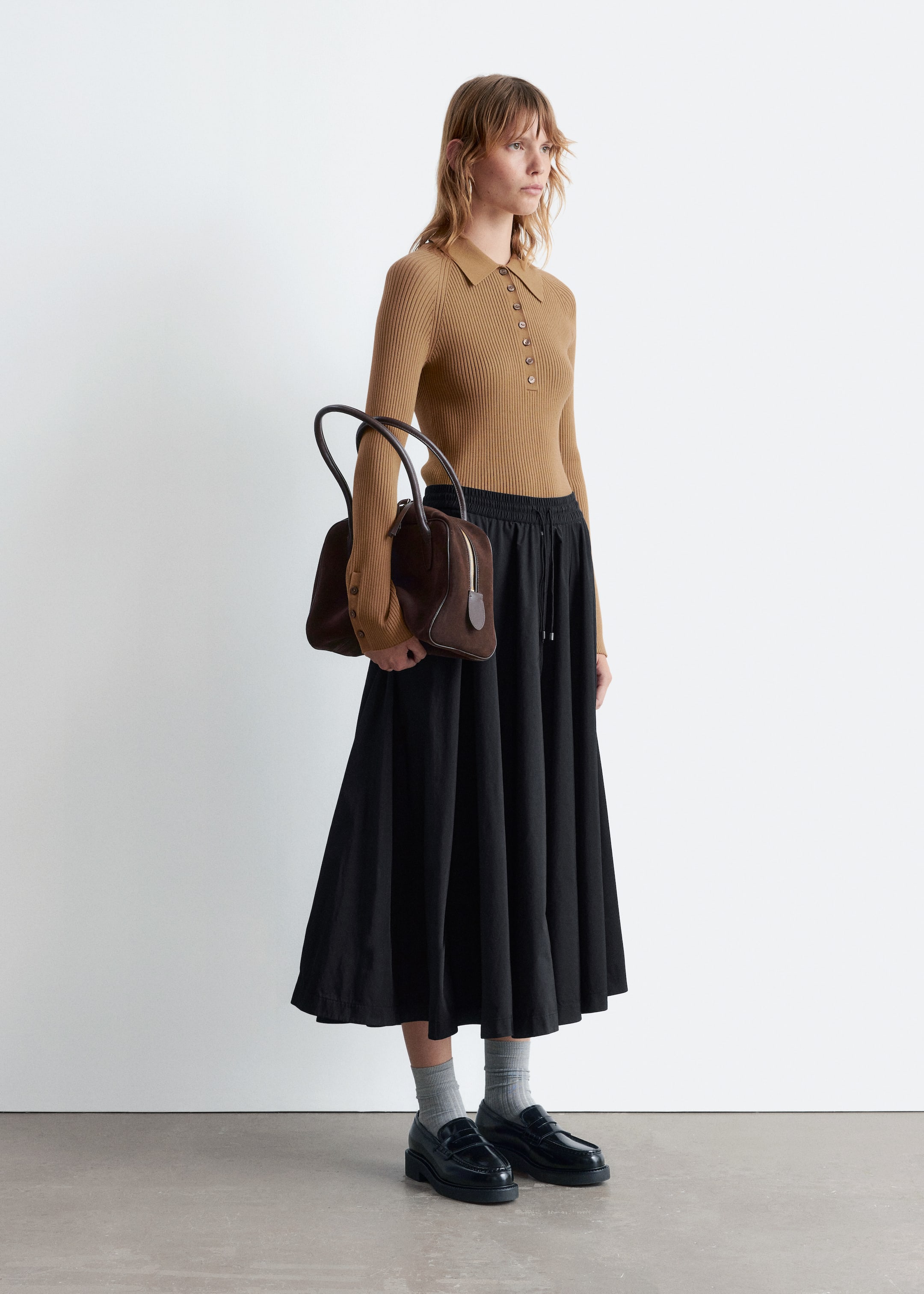 Agrandir l'image: Un polo à manches longues côtelé de couleur beige avec un col et une patte de boutonnage est porté, présenté en vue de profil, accompagné d'une jupe midi noire à taille élastique avec cordon de serrage. Des chaussettes grises côtelées et des mocassins noirs épais complètent le look, tandis qu'un sac à main structuré marron avec deux poignées supérieures est porté sur le côté.