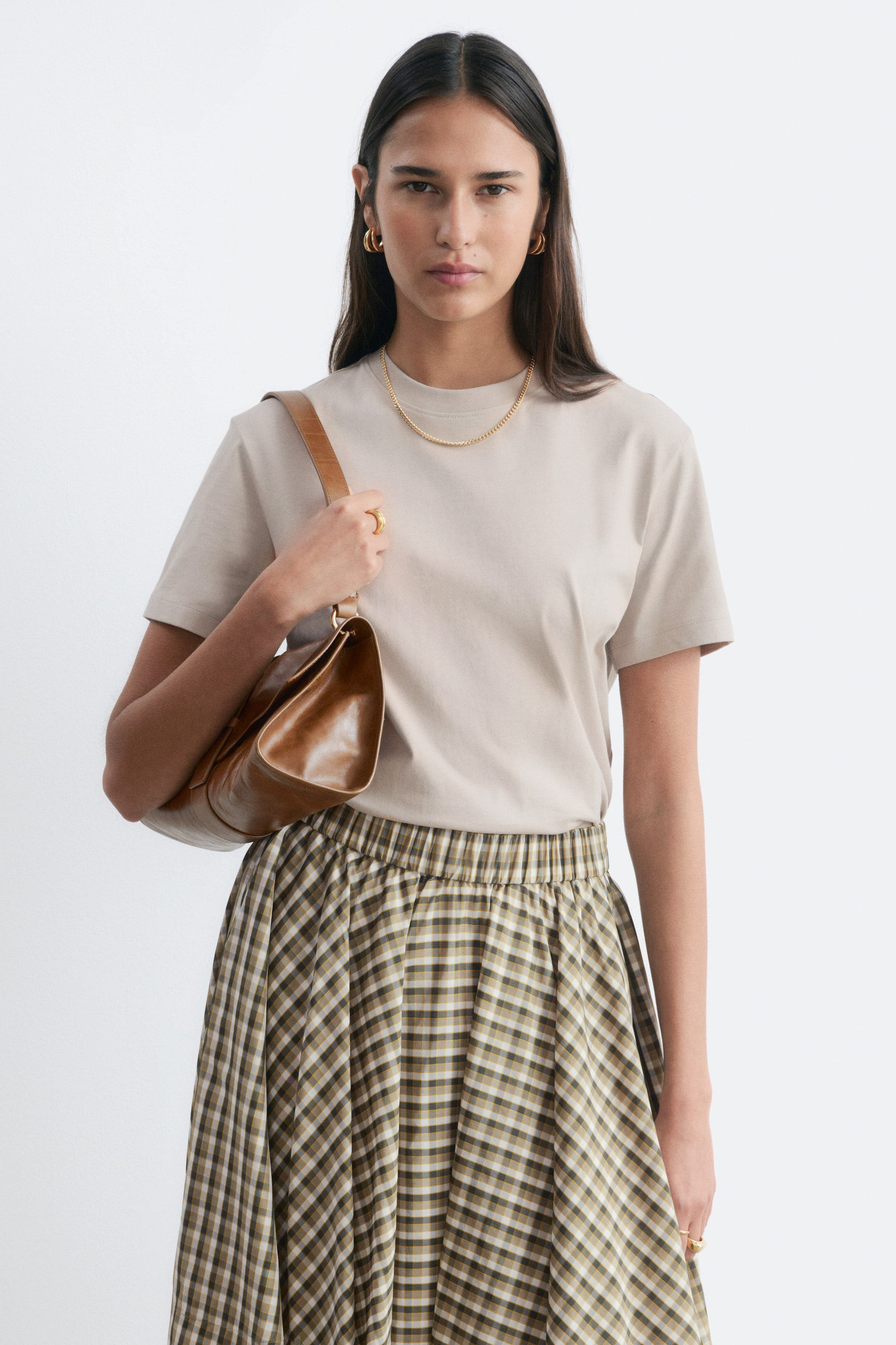 Grotere afbeelding bekijken: Een beige T-shirt met een normale pasvorm en een rechte snit, voorzien van een ronde halslijn en korte mouwen. Het model draagt het T-shirt in een geruite rok gestopt en gestyled met een gouden ketting.