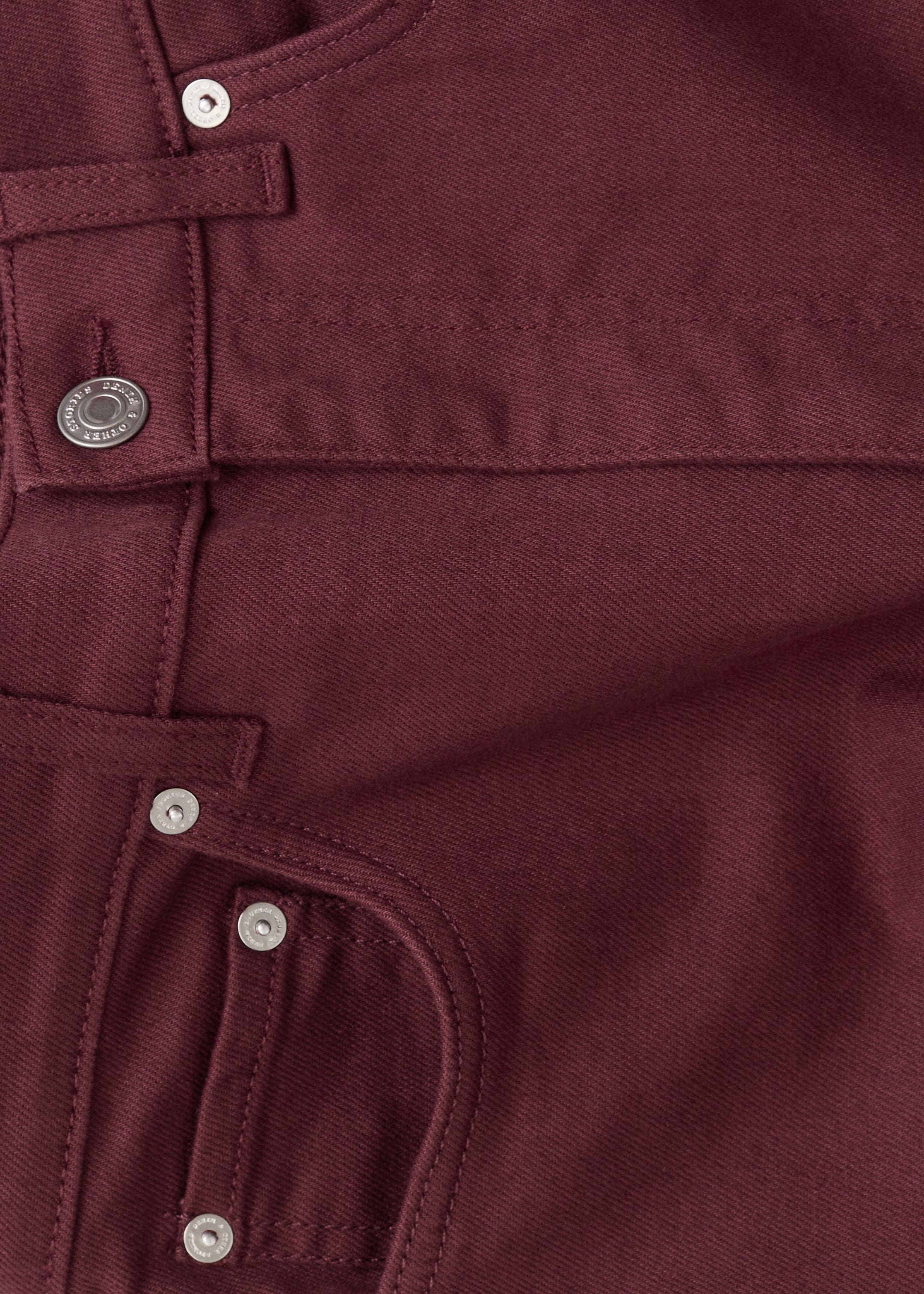 Ingrandisci l'immagine: La parte superiore dei jeans bordeaux è mostrata dall'alto, mostrando un tessuto denim a trama diagonale. Presenta due bottoni color argento, un passante per cintura e una tasca per monete con due rivetti color argento.