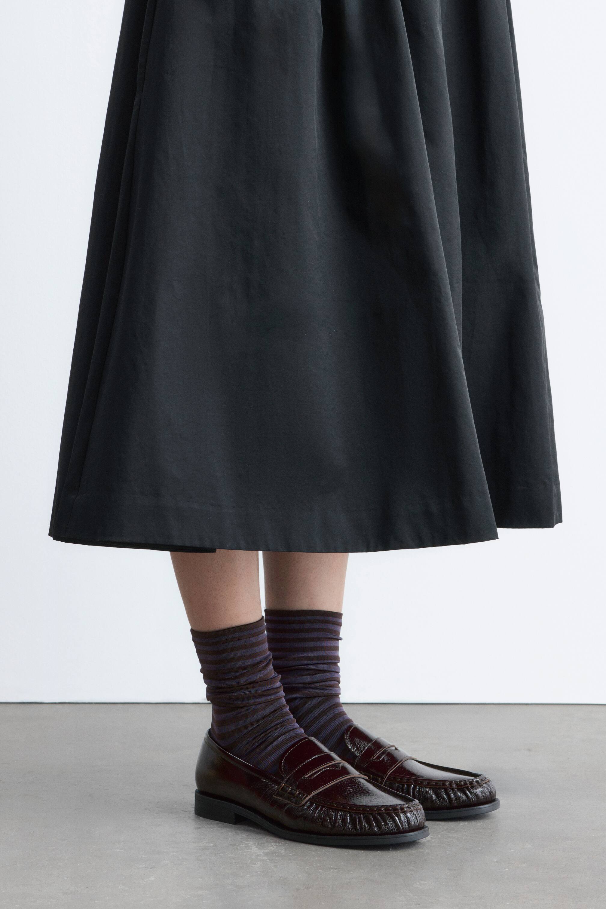 Ingrandisci l'immagine: Una gonna nera presenta una vita arricciata e una silhouette ampia e fluida, che cade a lunghezza midi. Il tessuto si drappeggia dolcemente attorno alle gambe, creando delicate pieghe verticali.