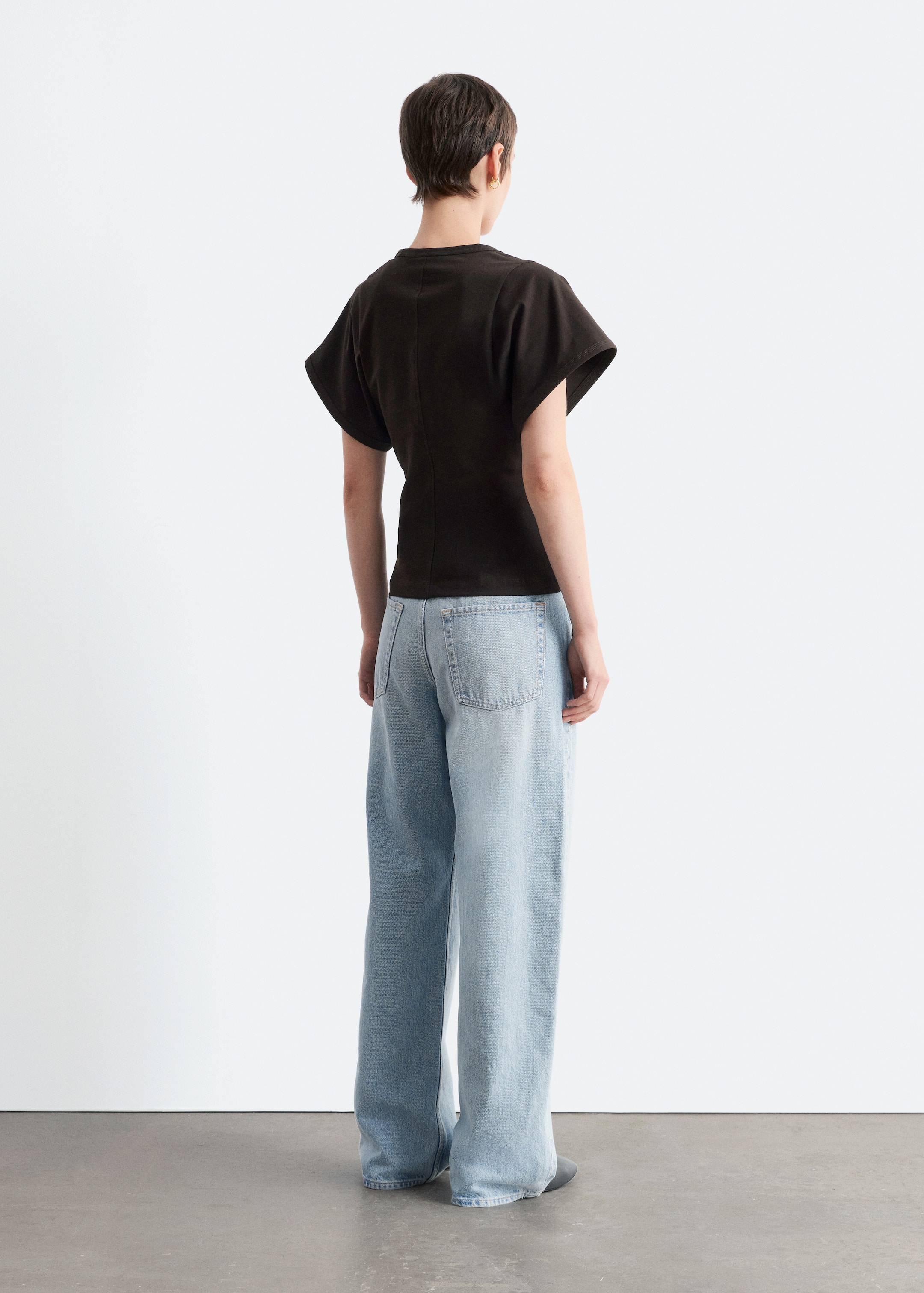 Grösseres Bild ansehen: Ein dunkles T-Shirt mit tailliertem Schnitt und kurzen, weiten Ärmeln wird vom Model getragen, das eine sichtbare, abgesteppt Naht entlang der hinteren Mitte zeigt. Das Model trägt auch hell gewaschene, weit geschnittene Jeans.