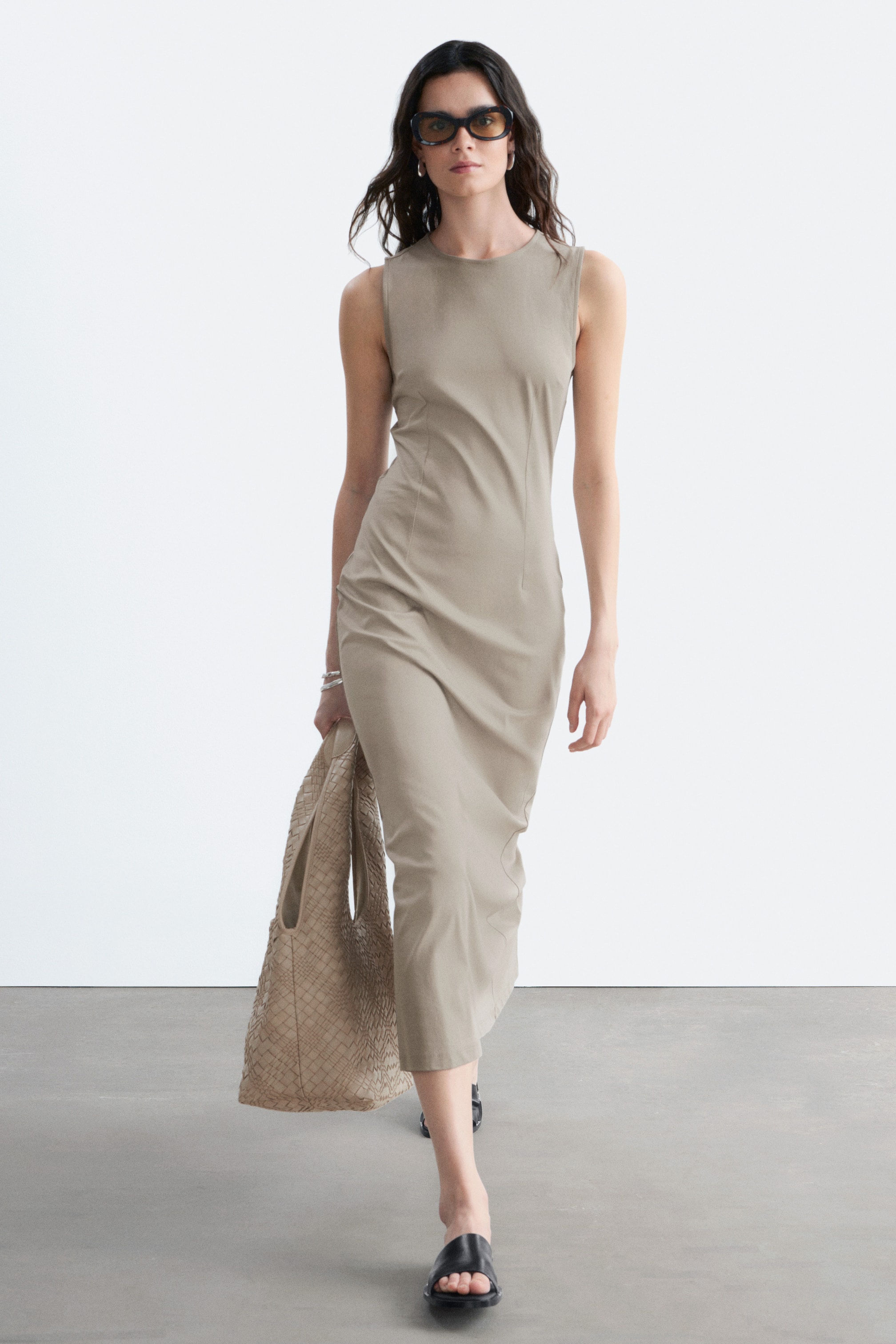 Agrandir l'image: Une robe midi sans manches de couleur beige clair est présentée sur un mannequin marchant légèrement en biais, mettant en valeur son encolure ras-du-cou, sa silhouette ajustée et ses détails de coutures verticales.