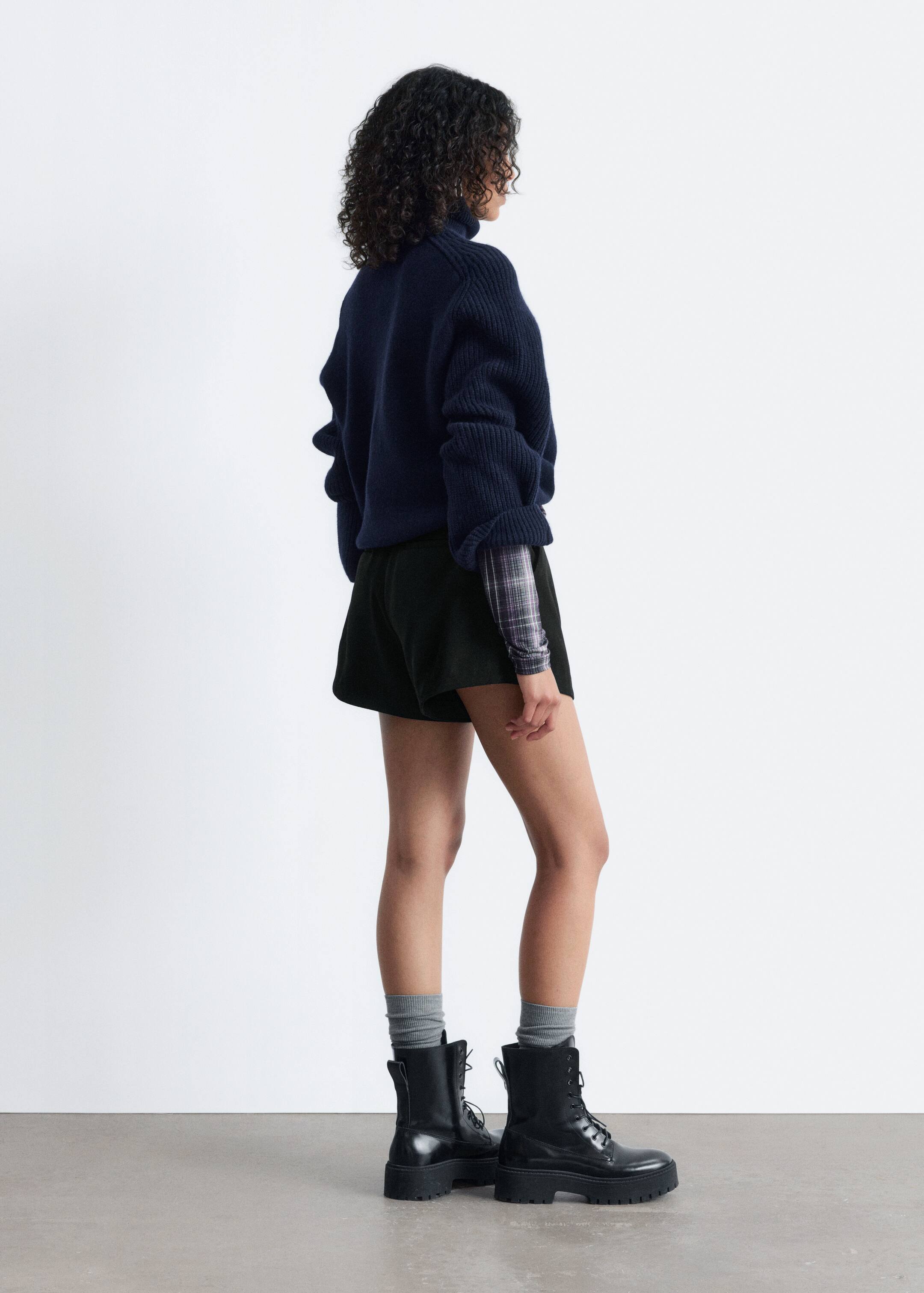 Visualizza immagine più grande: Un maglione dolcevita a maglia blu scuro con una texture a coste e una forma squadrata viene indossato dalla modella. Le sue maniche voluminose a coste si raccolgono ai polsi e una camicia a quadri è visibile stratificata sotto, estendendosi oltre l'orlo e i polsini del maglione.