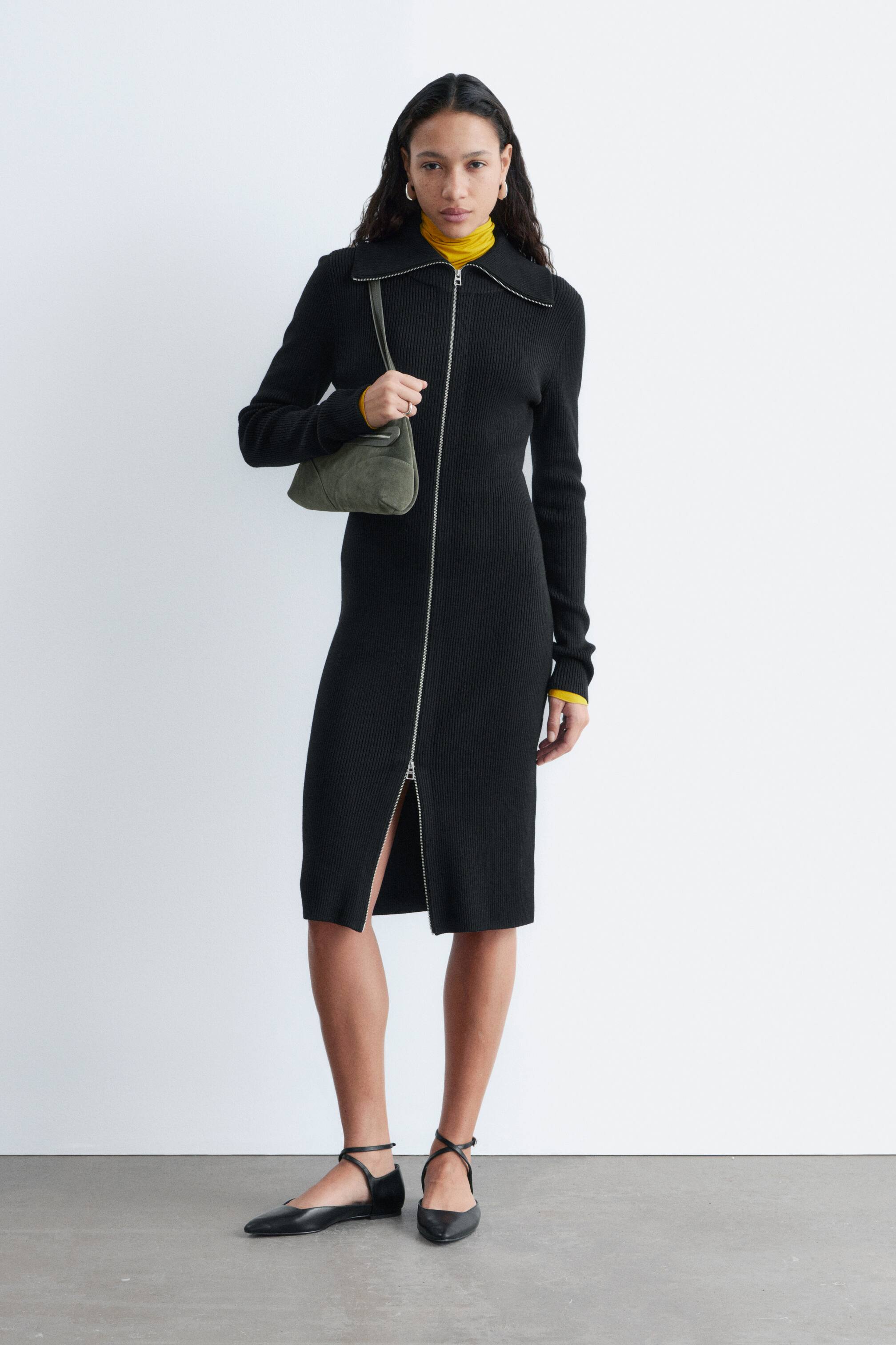 Ver imagen más grande: Un vestido midi negro de canalé, que presenta una silueta ajustada, mangas largas y un cuello doblado, es llevado por la modelo. Su cremallera plateada de doble sentido está parcialmente abierta desde la parte inferior, creando una abertura frontal, y un cuello alto amarillo se lleva debajo.