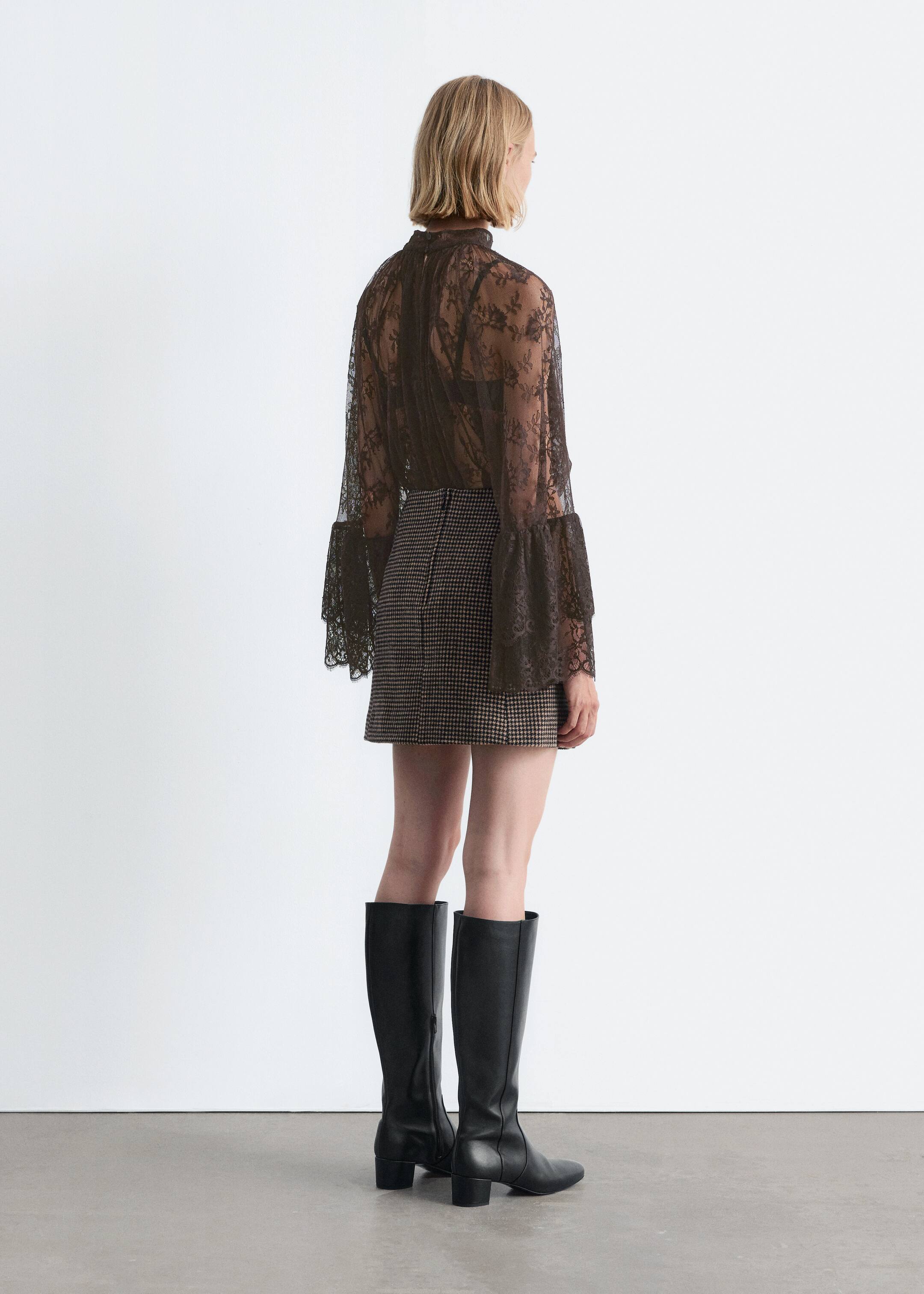View larger image: A dark, sheer lace top with a high neck and wide, flared cuffs is being shown from the back, layered over a dark bra and styled with a micro-houndstooth mini skirt and black knee-high boots with block heels.