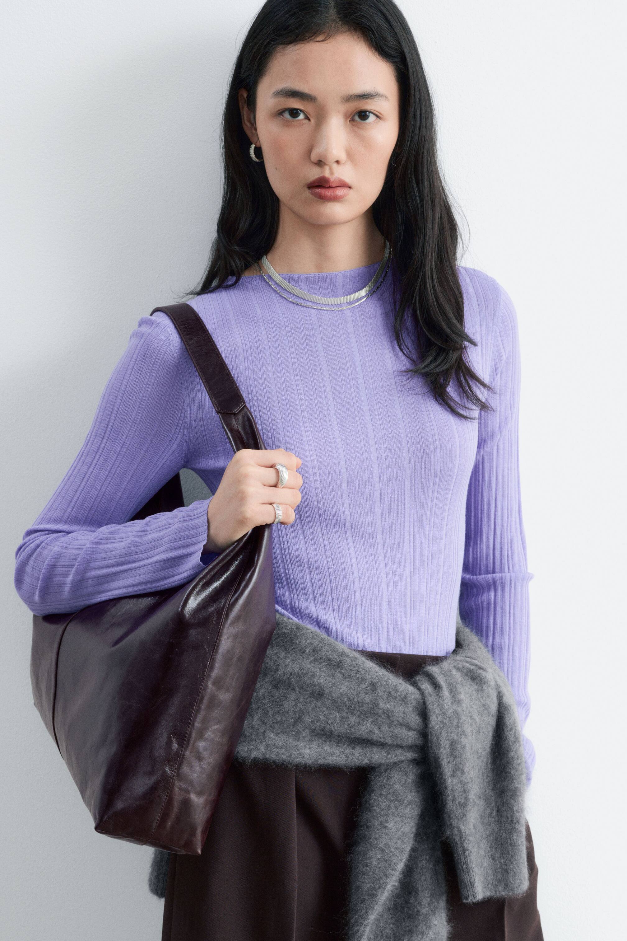 Ver imagen más grande: Un bolso tote grande y sin estructura, elaborado en piel de grano suave de color marrón oscuro, lo lleva la modelo colgado del hombro. Con la mano derecha sujeta el borde superior del bolso.