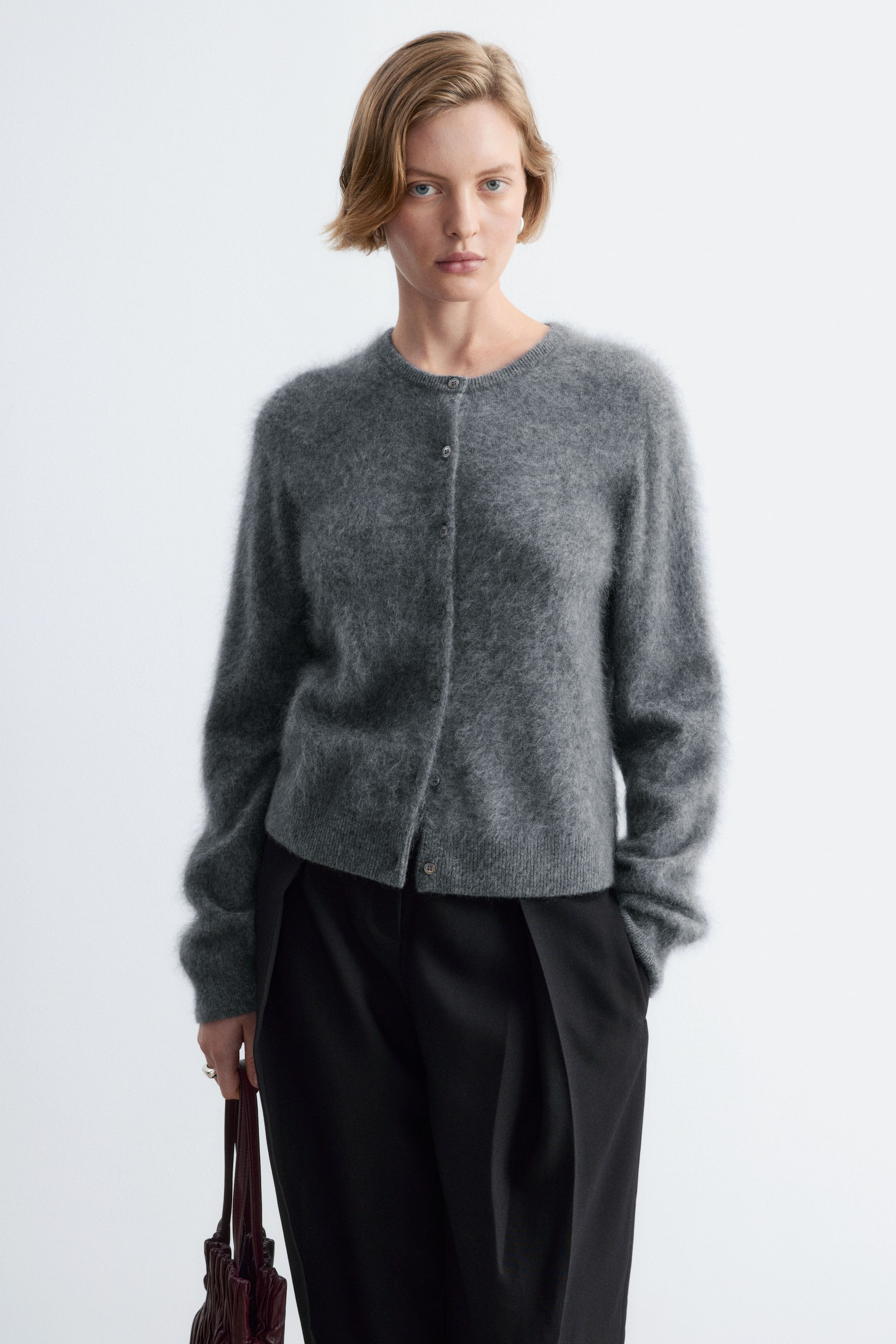 Agrandir l'image: Un cardigan en cachemire brossé gris, à la texture duveteuse, avec col rond et fermeture boutonnée sur le devant, est porté par le mannequin. Le cardigan est associé à un pantalon noir, et le mannequin tient un sac à main bordeaux foncé.