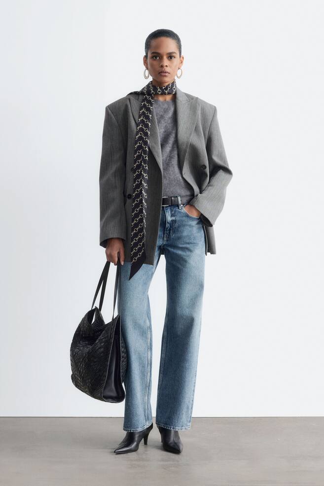 Una americana gris cruzada con solapas de muesca y corte relajado la lleva la modelo sobre un top gris y unos vaqueros azules. La americana se combina con un paÃ±uelo estampado drapeado alrededor del cuello y un bolso negro tejido. - 1
