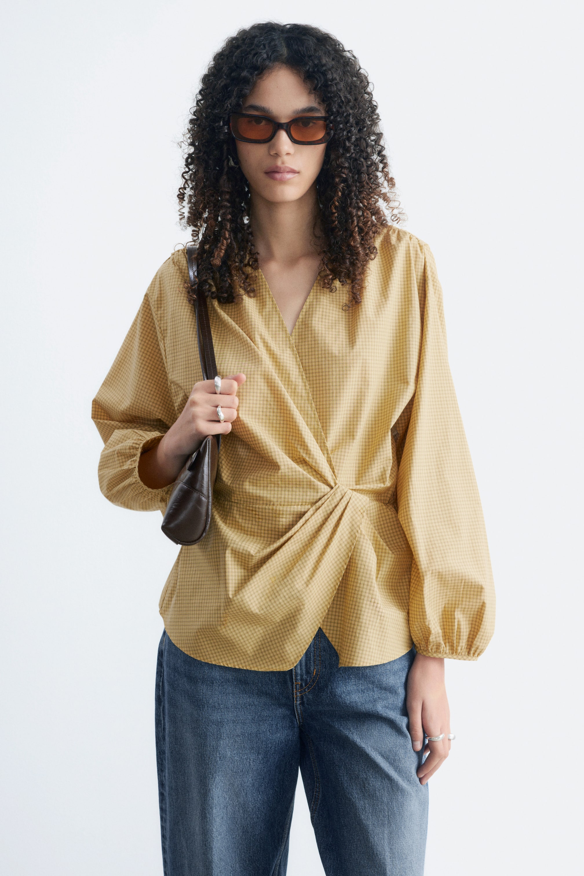 Agrandir l'image: Le mannequin porte une blouse cache-cœur en coton à motif quadrillé jaune. Elle présente un col V, des épaules froncées et des poignets froncés, ainsi qu’un détail marqué à la taille formant un faux nœud avec des plis. Le mannequin porte également un jean en denim bleu.
