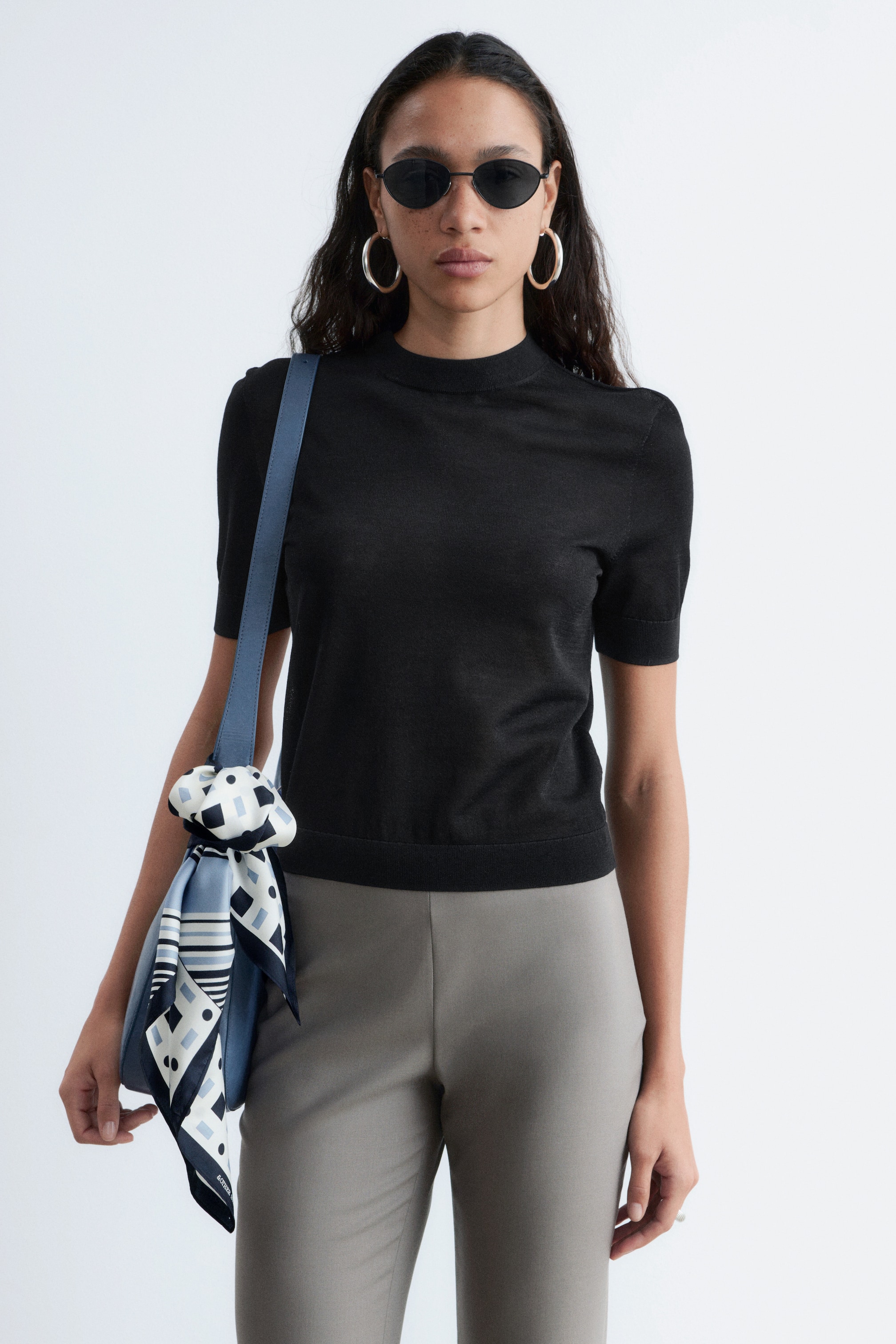 Ver imagem maior: A modelo está a usar uma T-shirt de malha preta, combinada com calças cinzentas. A T-shirt tem um corte ajustado com mangas curtas, gola redonda e acabamentos canelados na bainha e nas mangas, assentando na anca.