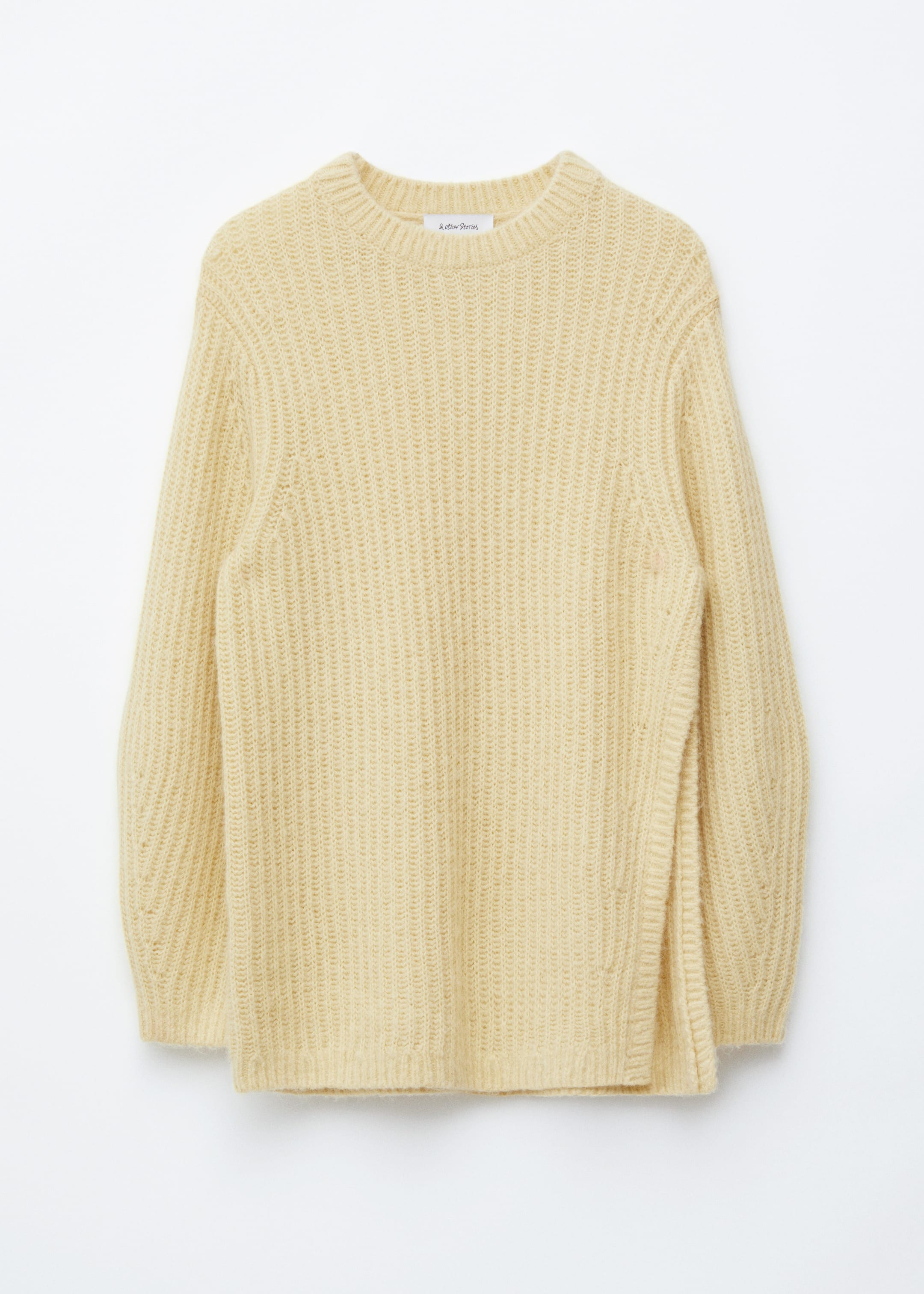 Visualizza immagine più grande: Un maglione lavorato a maglia di colore giallo chiaro è esposto rivolto verso il FRONTE, caratterizzato da uno scollo rotondo, bordi a coste e una silhouette complessivamente oversize e lunga. Presenta maniche lunghe e voluminose e uno spacco alto sul lato destro dell'orlo, con un evidente motivo a coste verticali su tutta la superficie.