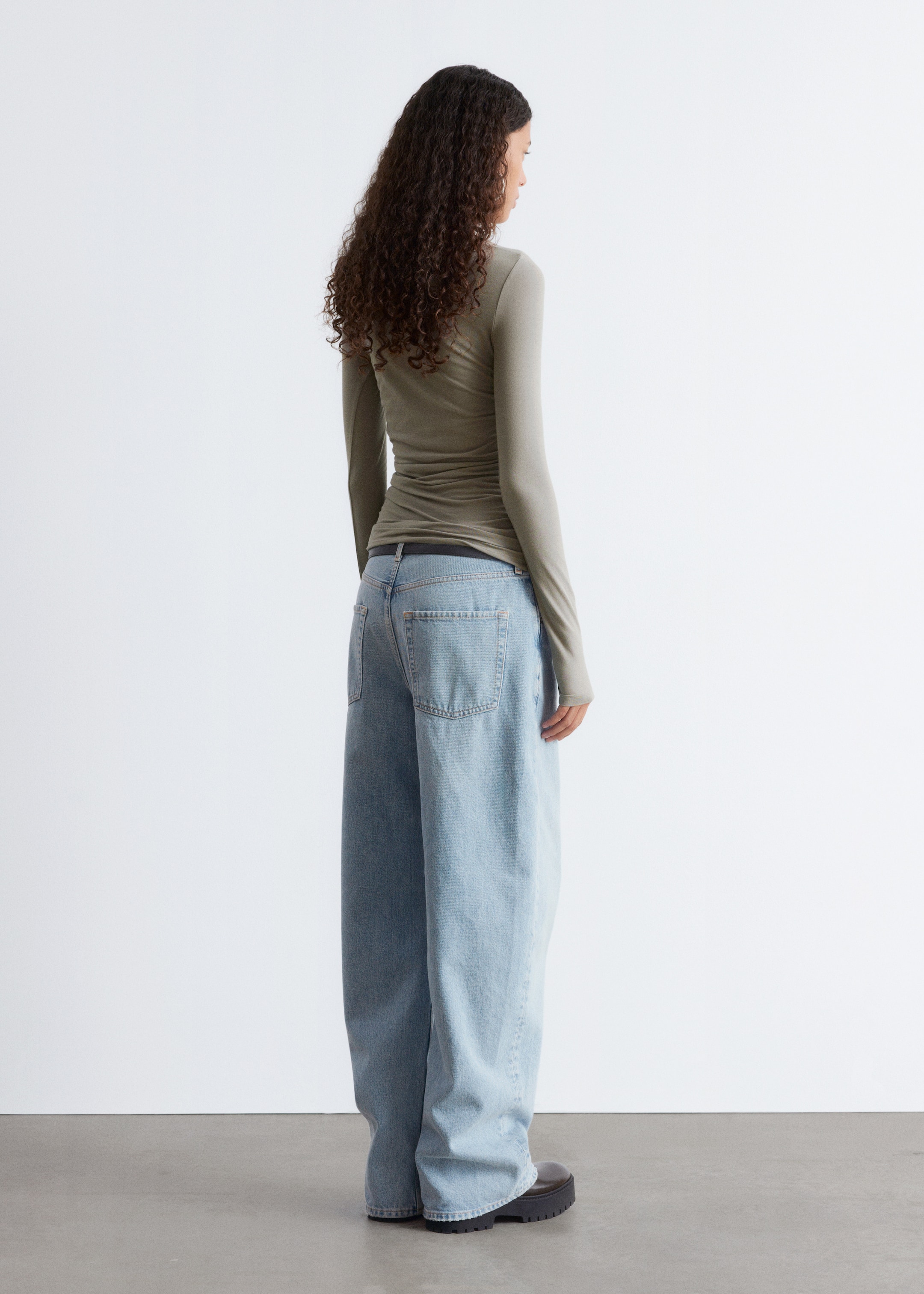 Visa större bild: En olivgrön långärmad topp presenteras från baksidan till höger, den sitter tätt och har rynkningar runt midjan. Ljus tvätt, vida jeans har synliga bakfickor och en avslappnad passform, som täcker svarta skor med chunky-sulor.