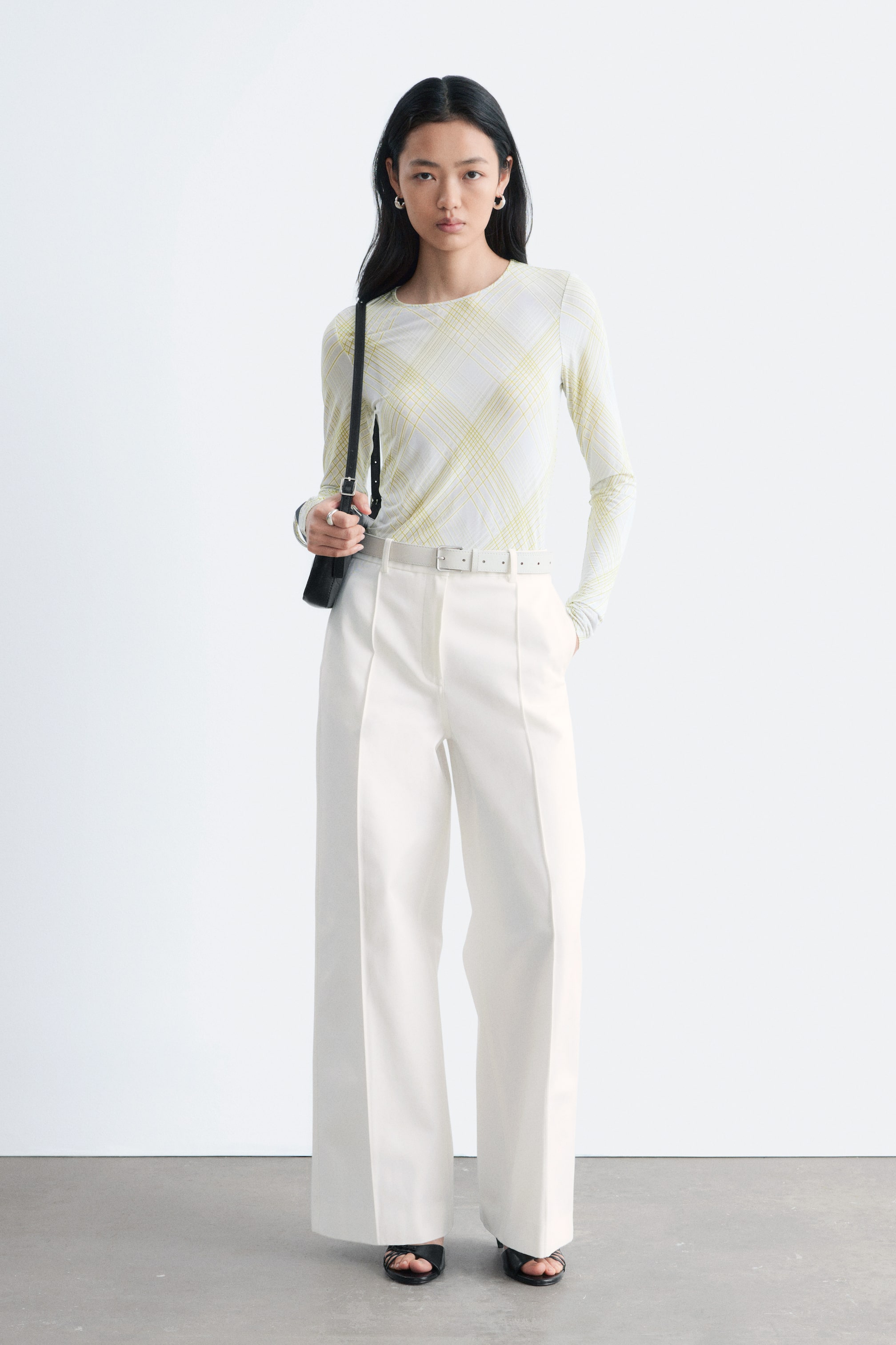 Προβολή μεγαλύτερης εικόνας: Ένα μακρυμάνικο μπλουζάκι από ζέρσεϊ με σχέδιο καρό σε ανοιχτό κίτρινο και στρογγυλή λαιμόκοψη φοριέται από το μοντέλο. Διαθέτει ίσια σιλουέτα και συνδυάζεται με λευκό παντελόνι με φαρδιά μπατζάκια και λευκή ζώνη.