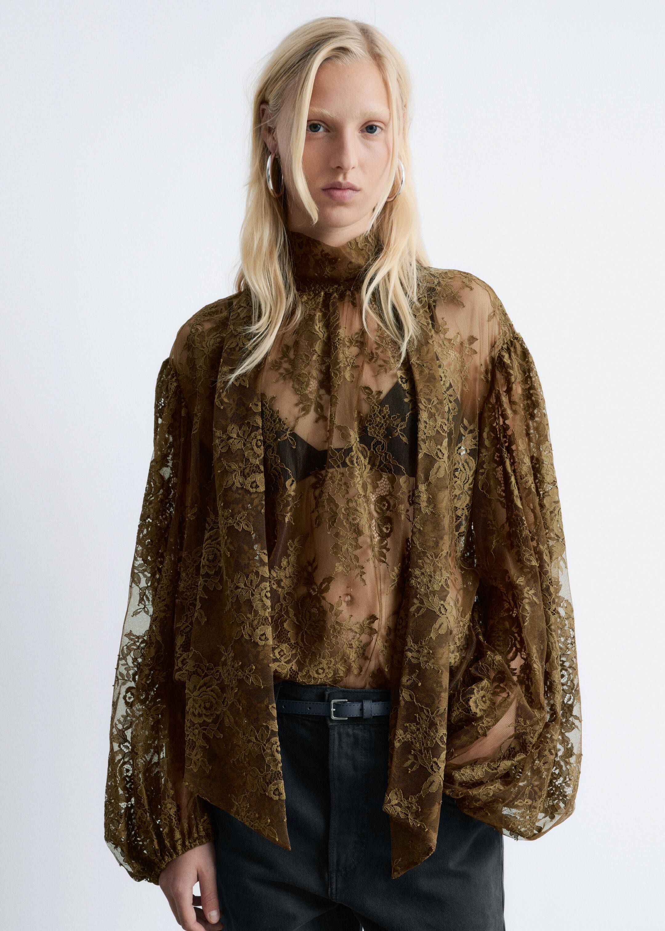 Ingrandisci l'immagine: La blusa di pizzo floreale trasparente verde oliva viene presentata frontalmente, con un collo alto e maniche lunghe e voluminose che si raccolgono nei polsini. Sotto si vede un reggiseno nero e la blusa viene indossata sopra pantaloni neri con cintura nera.
