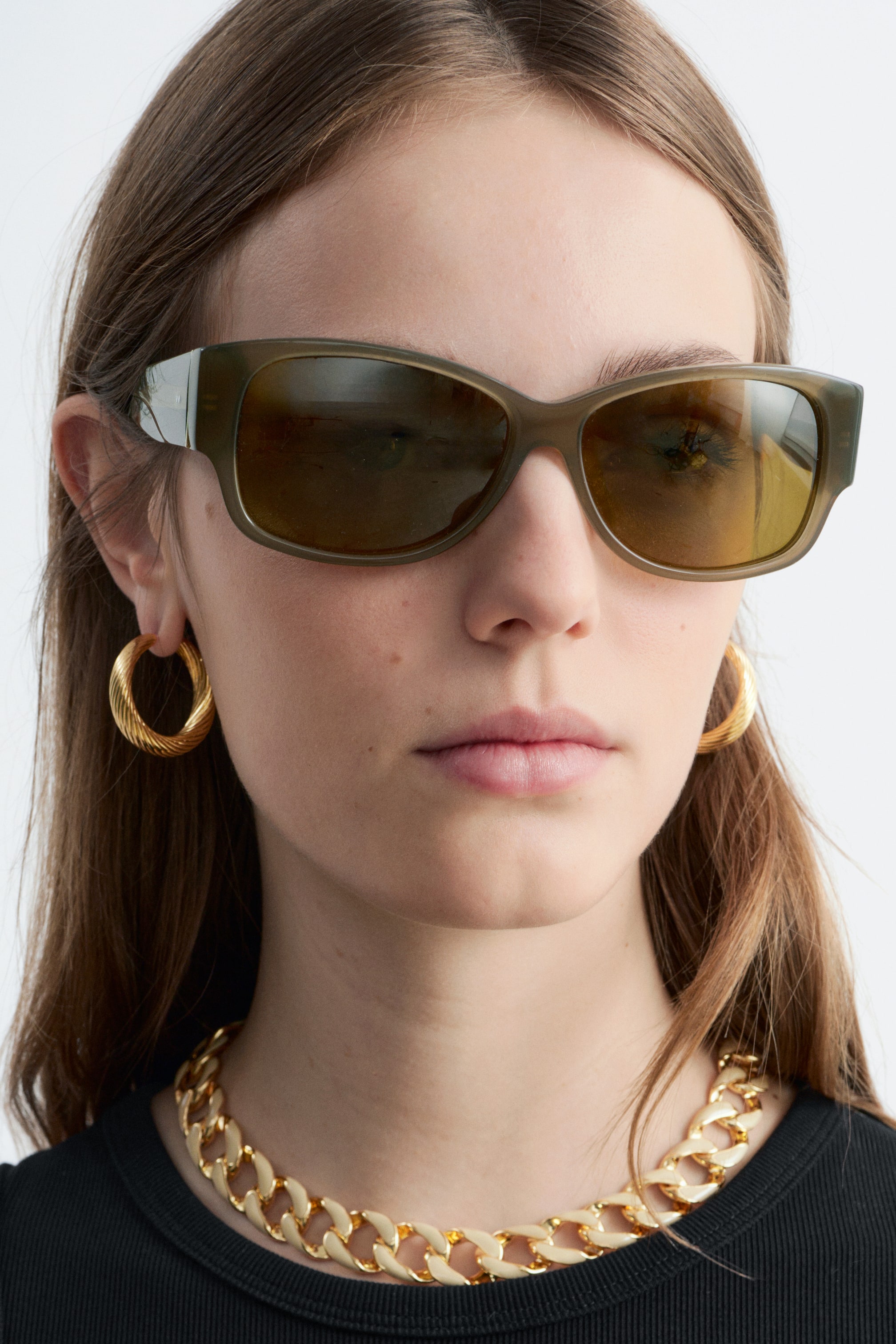 Se større billede: Oliven grønne rektangulære solbriller bæres, med mørke tonede linser og tykke stænger, der inkluderer en kontrasterende lysere detalje nær hængslet.