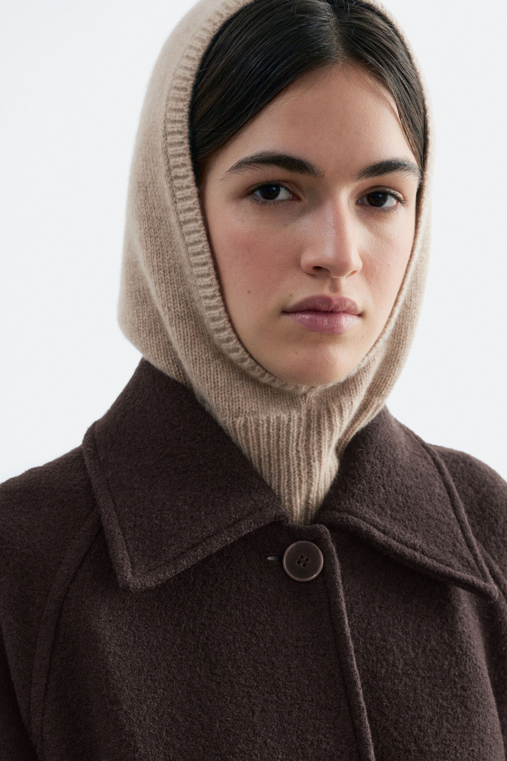 Visualizza immagine più grande: Un passamontagna con cappuccio in maglia beige e bordo a coste è indossato dalla modella, incorniciando il viso e coprendo il collo. Il cappuccio è indossato sotto un cappotto testurizzato marrone scuro con ampio colletto.