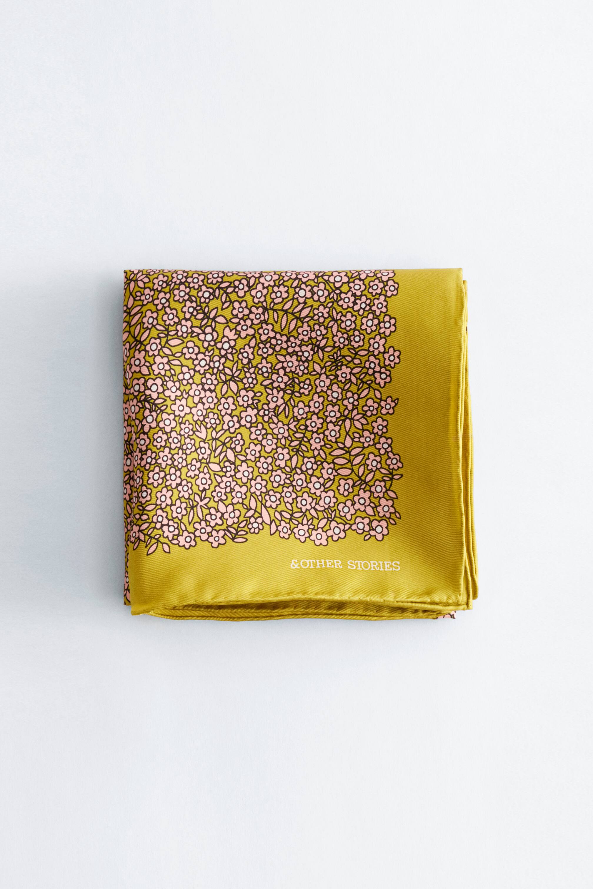 Se større billede: Et kvadratisk tørklæde er foldet fladt og viser sin forside. Det har en glansfuld olivengrøn stof med et tæt blomster mønster af små lyserøde blomster og sorte blade, der dækker den øverste venstre del. Mærkenavnet '& OTHER STORIES' er synligt i hvidt på det ensfarvede olivengrønne område.