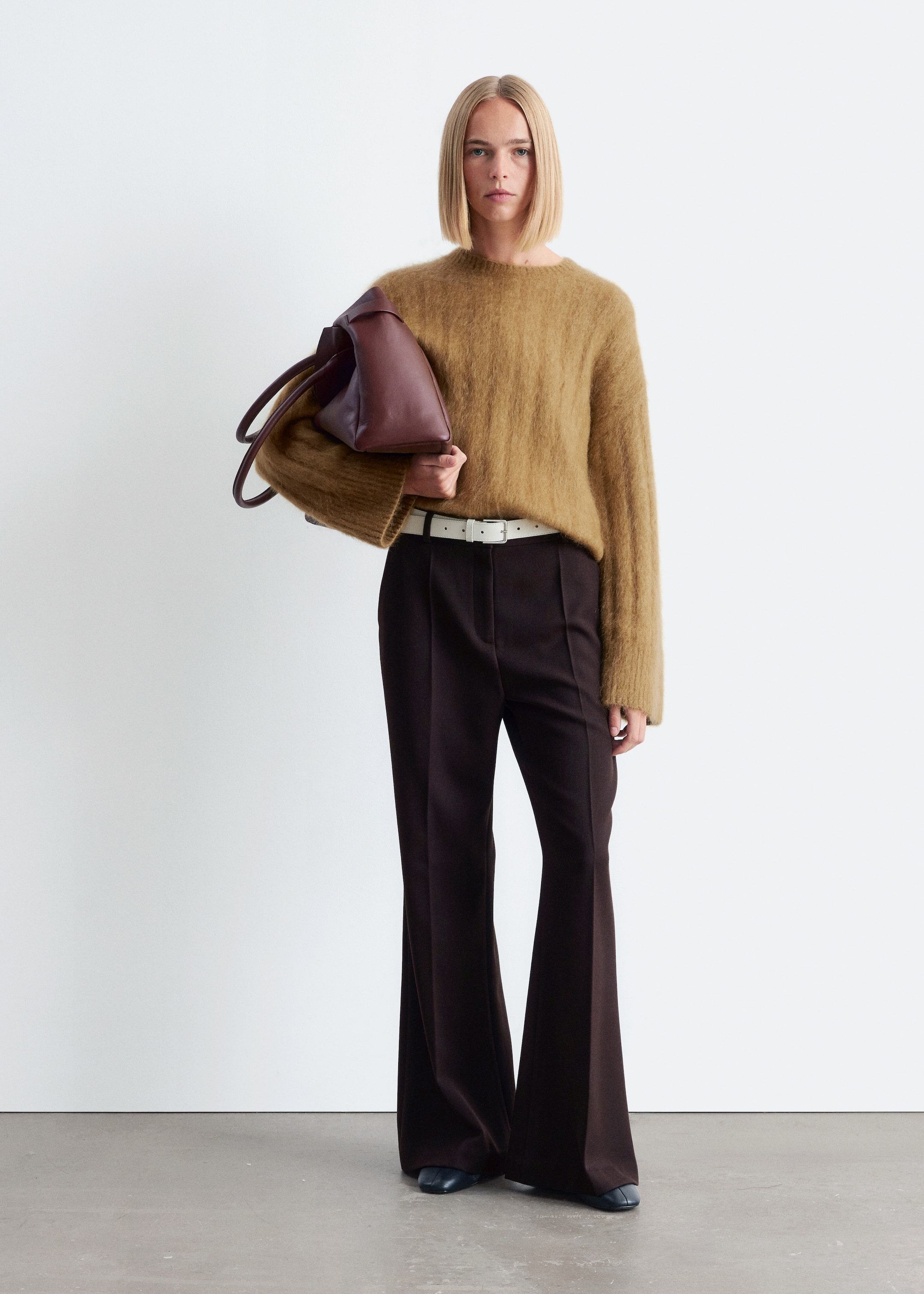 Agrandir l'image: Un pull en tricot duveteux brun-tan présente une coupe décontractée et des poignets larges côtelés, et est porté sur un pantalon évasé taille haute brun foncé qui met en valeur des plis avant prononcés. Une fine ceinture blanc cassé serre le pantalon, et un sac fourre-tout bordeaux non structuré est tenu, tandis que des chaussures plates foncées sont visibles.