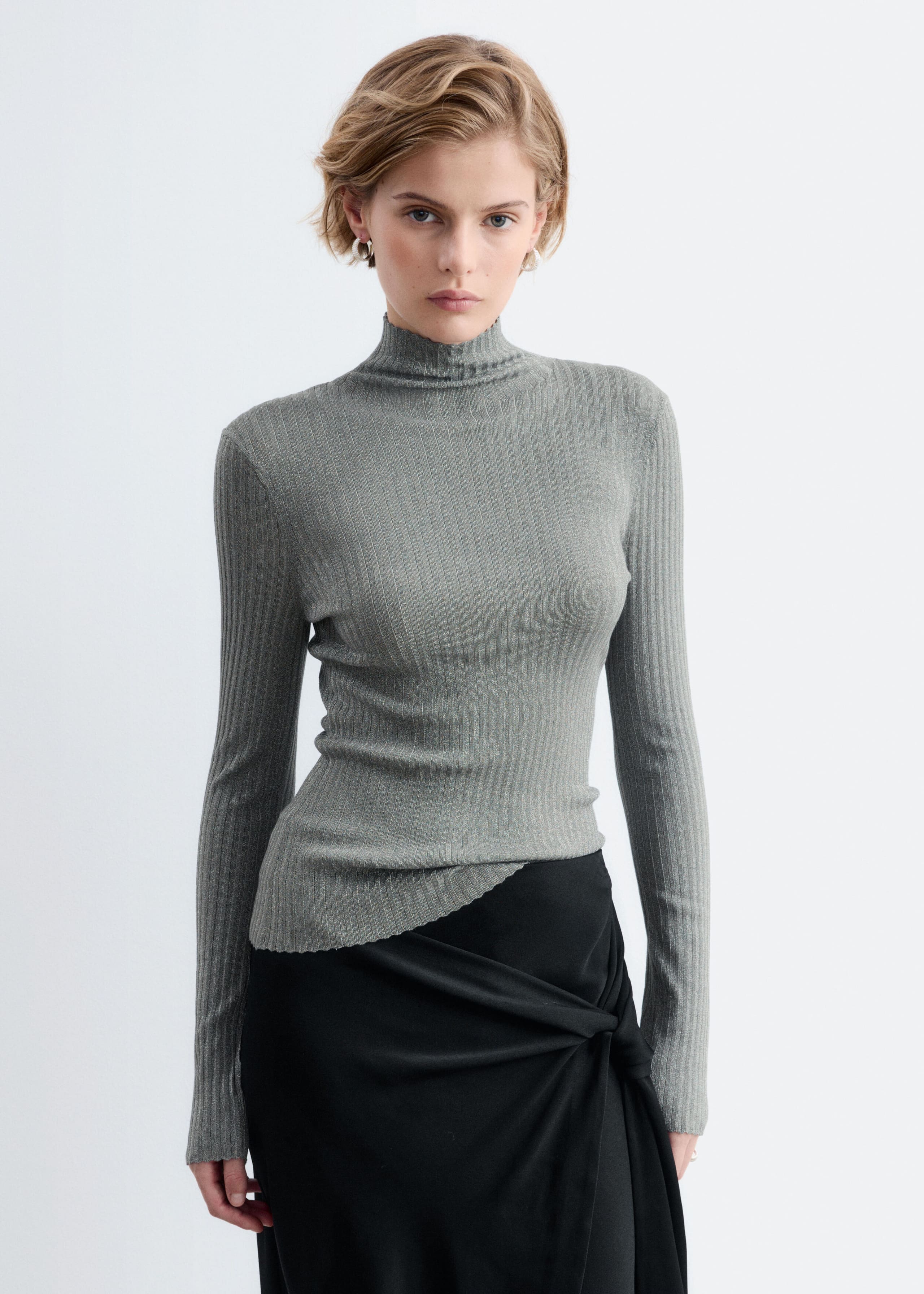 A gray rib-knit top with a subtle metallic sheen and a mock neck is being worn by the model. The long-sleeved top features a scalloped hem and is paired with a draped black skirt.