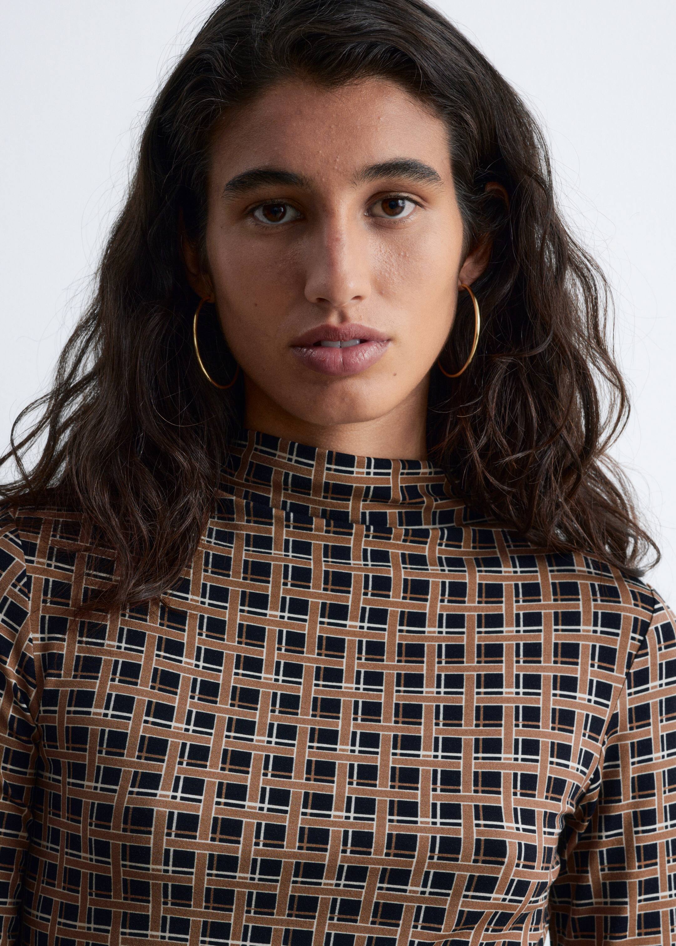 Größeres Bild ansehen: Ein Mock-Neck-Top wird getragen, das ein geometrisches Muster aus ineinandergreifenden braunen und schwarzen rechteckigen Formen aufweist, die mit dünnen weißen Linien umrandet sind. Das Top liegt eng um den Hals und die Schultern an und präsentiert so sein Design.